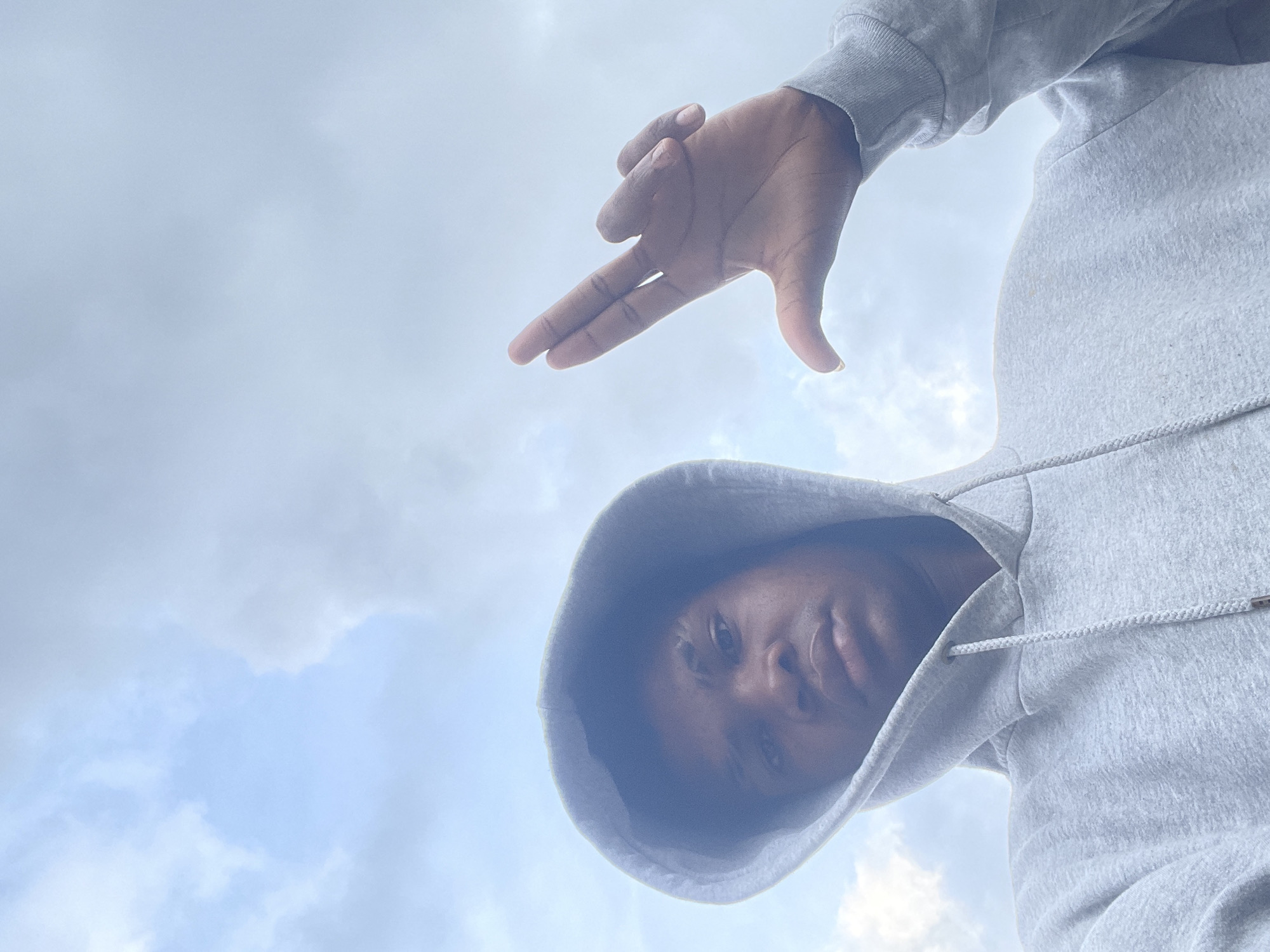 A low-angle photograph captures a young individual, appearing male, against an overcast sky in Ado, Nigeria. The subject, with dark skin, is wearing a heather grey hooded sweatshirt with the hood pulled over their head, casting shadows on their face. The individual looks directly into the camera with a neutral expression. Their right hand is raised, making a distinct gesture where the index and middle fingers are extended straight and together, the thumb is extended downwards, and the ring and pinky fingers are curled into the palm, forming a shape resembling the letter 'L'. The background is entirely sky, featuring a mix of light blue areas and diffused white and grey clouds, characteristic of an outdoor, daytime environment.