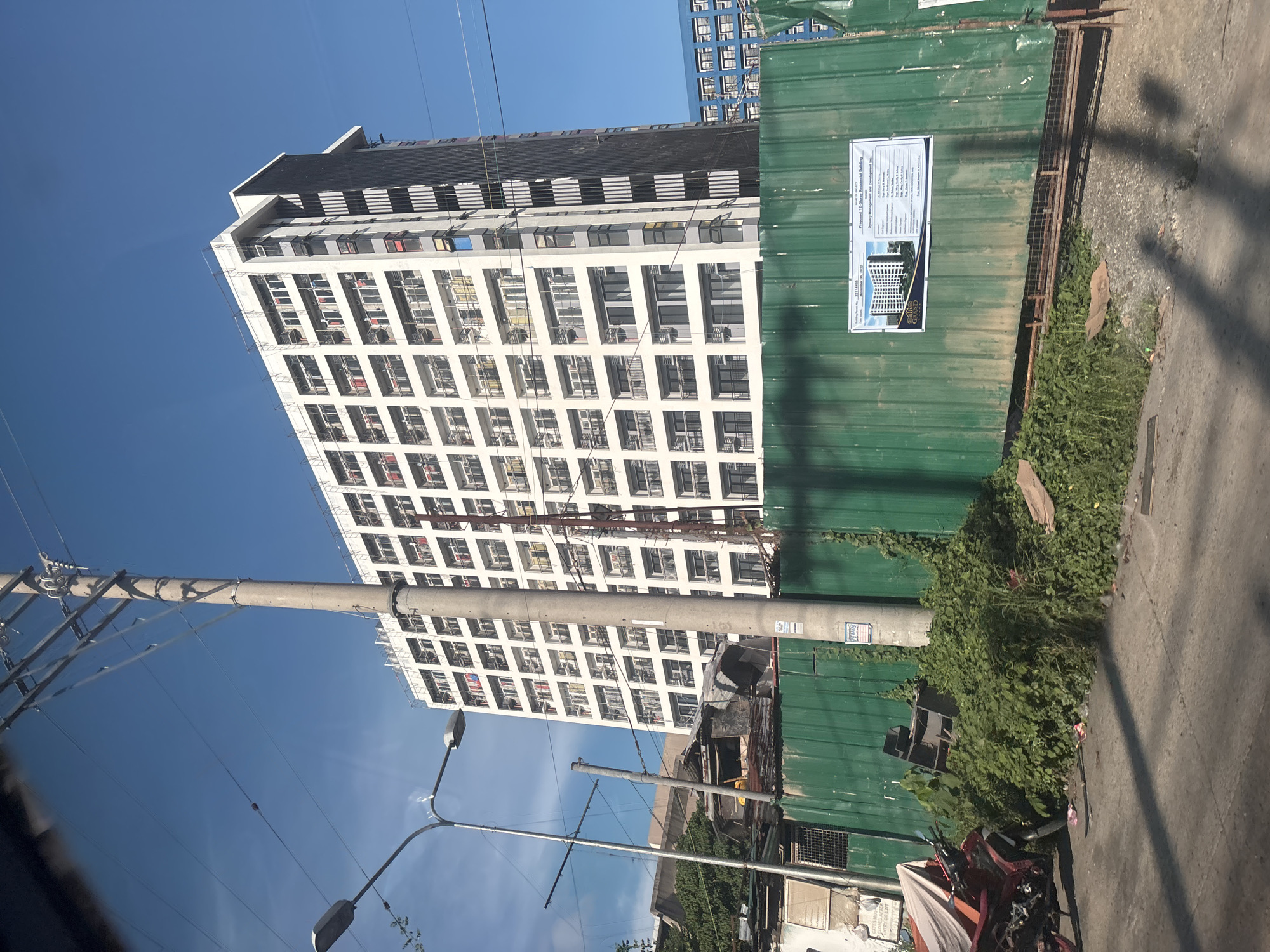 The image depicts a construction site in Davao City, Philippines, dominated by a tall, modern residential building under construction. The building is white with numerous balconies and windows, and is partially obscured by a tall, concrete utility pole. In front of the building, a green corrugated metal fence separates the construction area from the street. Overgrown vegetation spills over the fence in some areas, and a red motorcycle is parked to the left of the fence. A sign on the fence displays an architectural rendering of the completed building and lists project details. The sky is a clear, bright blue, suggesting a sunny day. The overall scene captures a moment of urban development, with the juxtaposition of the new construction against the existing urban landscape.