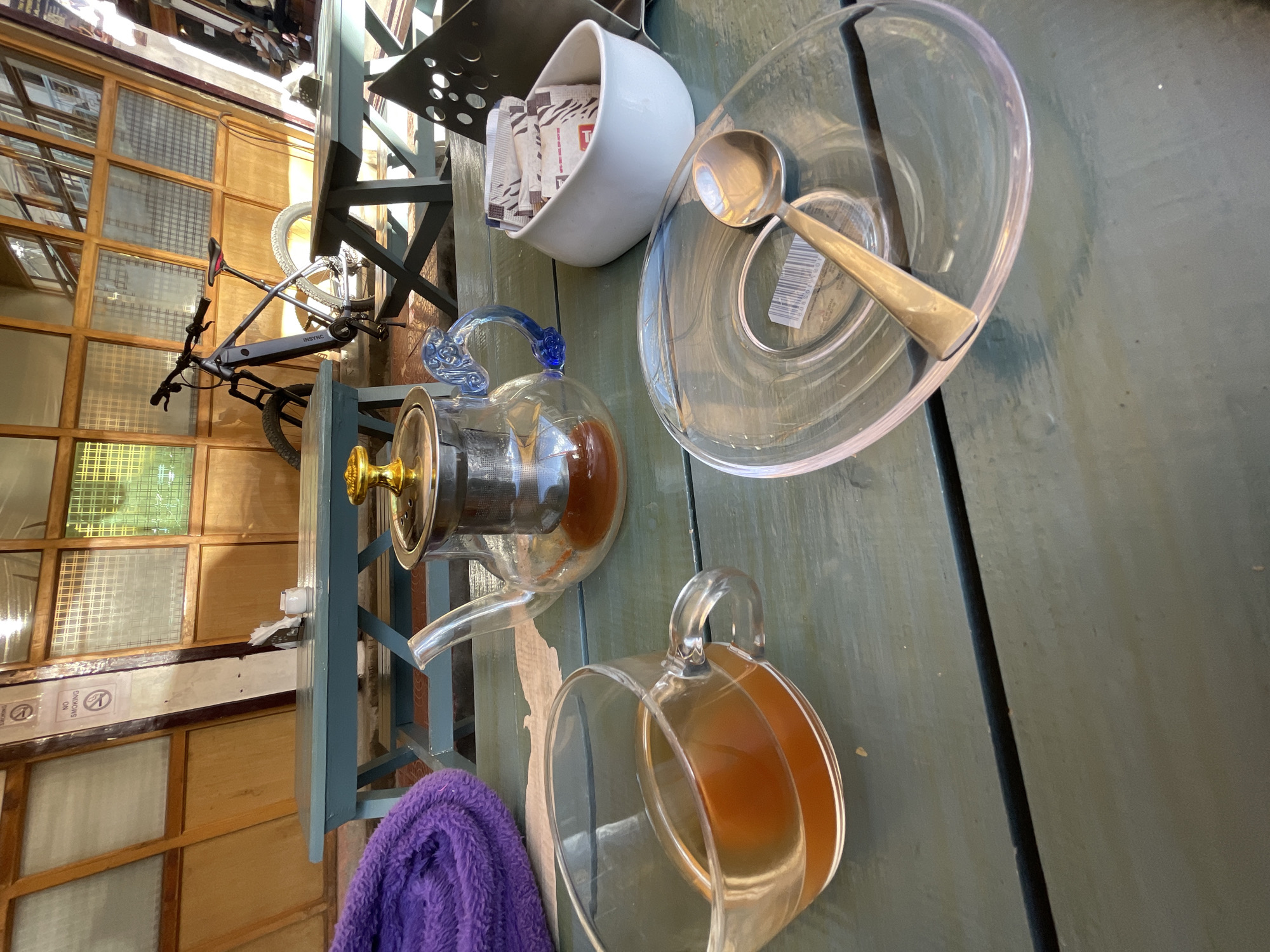The image shows a close-up view of a table setting at what appears to be an outdoor cafe or restaurant. The table is made of weathered, light green wooden planks. In the foreground, to the left, is a clear glass mug filled with amber-colored liquid, likely tea. Next to it is a transparent glass teapot with a silver infuser, also containing tea. The teapot has a decorative blue glass handle and a gold lid. To the right of the teapot is a small white ceramic bowl filled with several individually packaged sugar or sweetener packets. A silver spoon rests on a clear glass saucer in the lower right portion of the frame. A purple, folded towel or blanket is visible on the far left. In the background, a bicycle with a dark frame is parked near a table. Behind the table, there is a wooden structure with multi-paned windows, some of which have a grid pattern. Two "No Smoking" signs are visible on a wooden post in the background. The overall impression is a relaxed, casual setting. The presence of "No Smoking" signs and the architectural style with wooden elements suggests this could be a location in Leh, India, where such elements are common in cafes and guesthouses.