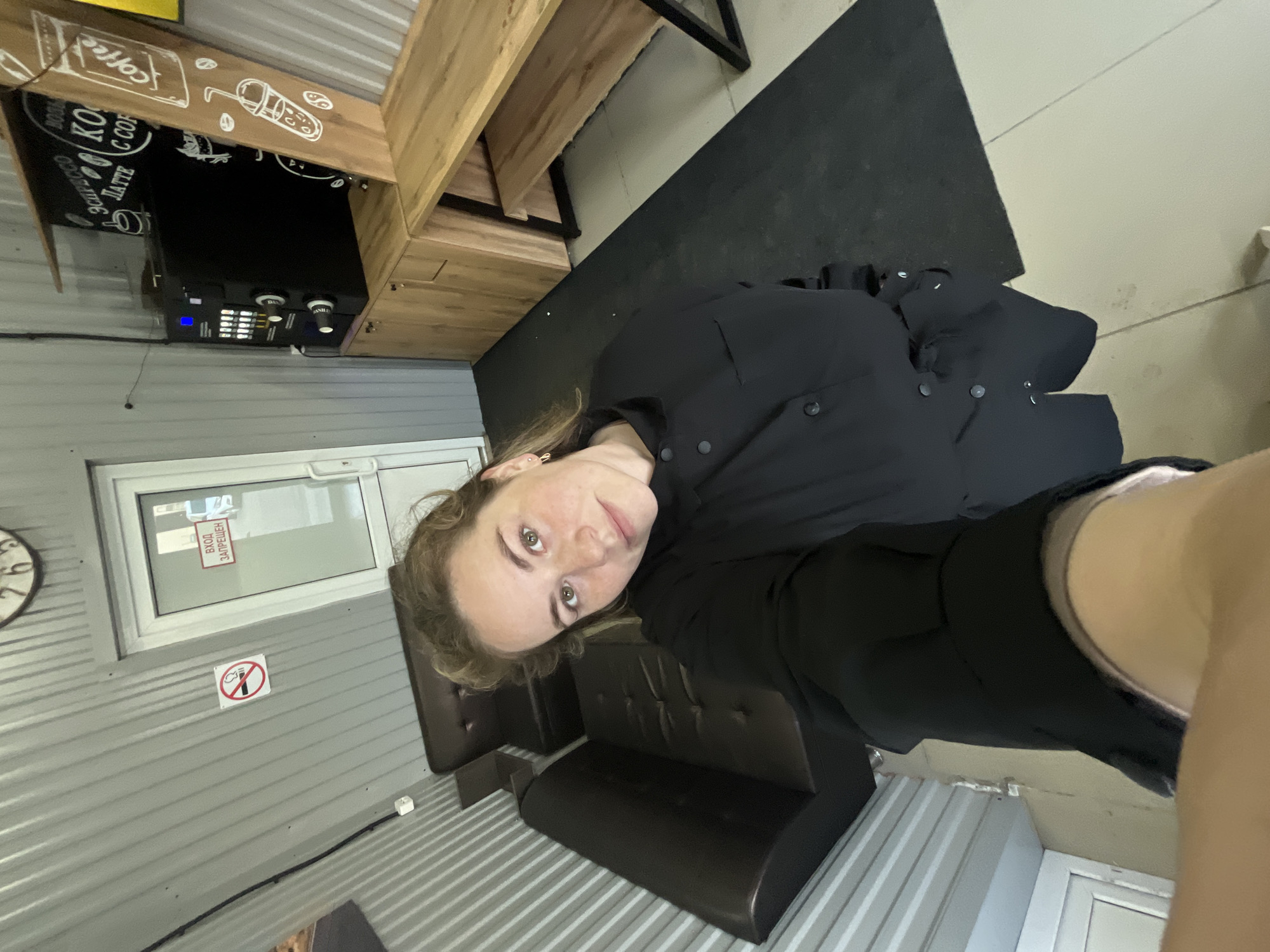 A selfie photograph taken inside a building, possibly a cafe, in Chaikovsky, Russia. A fair-skinned individual with blonde hair is the primary subject. The person is wearing a black button-down shirt or jacket. The background includes a corrugated metal wall, a window with a sign in Russian that says "Entrance Prohibited," and a "No Smoking" sign. There is also a dark leather bench or seating area in the background. Visible towards the upper-left of the frame is a coffee machine and branding, suggesting a cafe or coffee shop setting. Additional furniture or fixtures can be seen in the upper portion of the image.