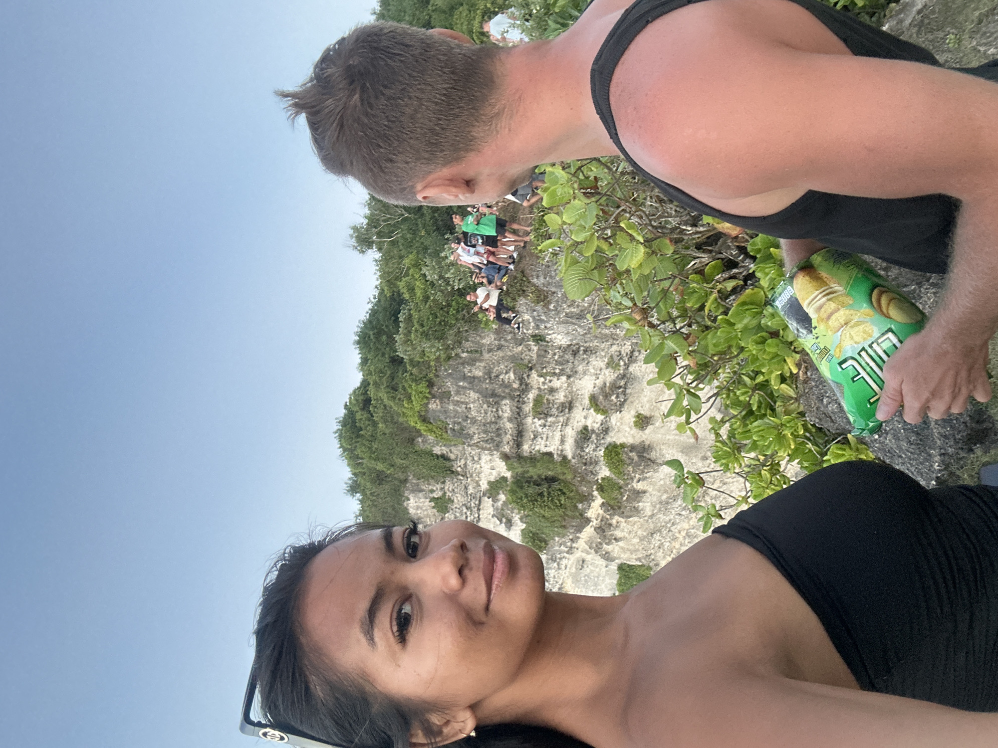 In Kuta Selatan, Indonesia, a young woman with dark hair and sunglasses on her head is taking a selfie. She wears a black strapless top and smiles at the camera. Behind her, a man in a black tank top is partially visible, holding a can of chips. They are standing on a rocky cliff overlooking the ocean, with lush greenery and a sheer cliff face behind them. Several other people are gathered on the cliff in the background, observing the view. The sky is clear and blue.