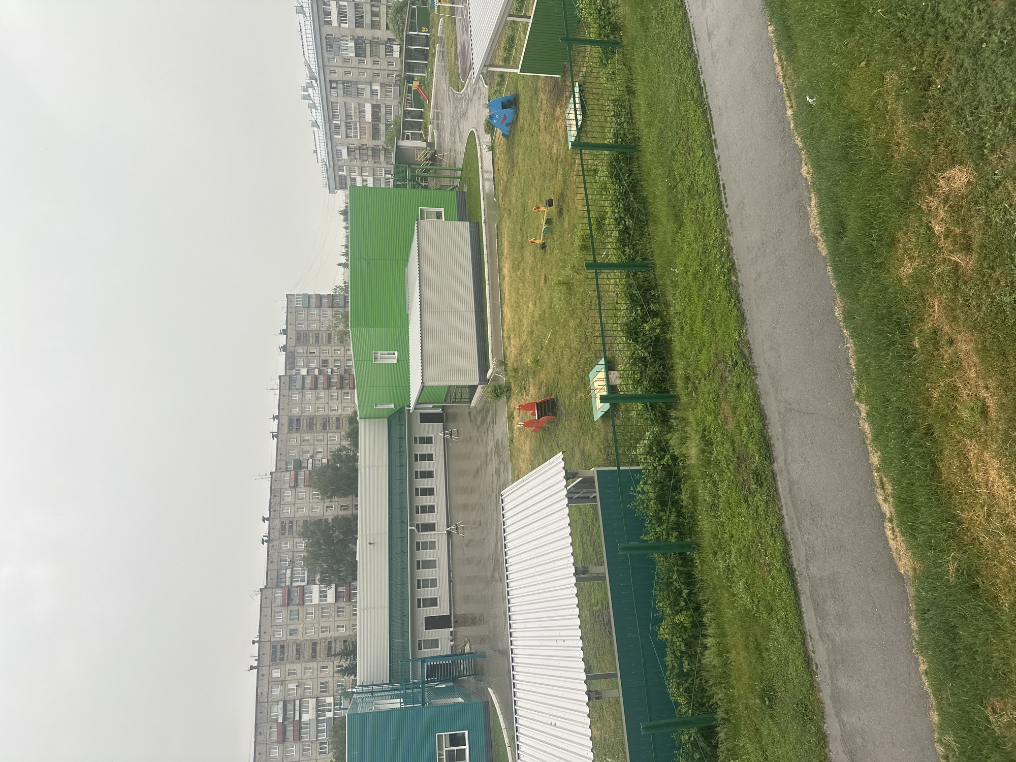 The image captures an overcast day in Novoaltaysk, Russia, showcasing a section of a school or kindergarten courtyard. In the background, multiple Soviet-era apartment buildings with similar architectural styles and muted color palettes dominate the skyline. The foreground features a children's play area with various equipment including a red slide structure, a blue dolphin-shaped climbing toy, a small seesaw, and a sandpit covered by a metal roof. A low green fence encloses the play area and separates it from a paved walkway and grassy areas. Adjacent to the play area is a modern, low-rise building with a green roof and siding, likely part of the educational facility. The ground is wet, suggesting recent rain. There are no people visible in the scene.