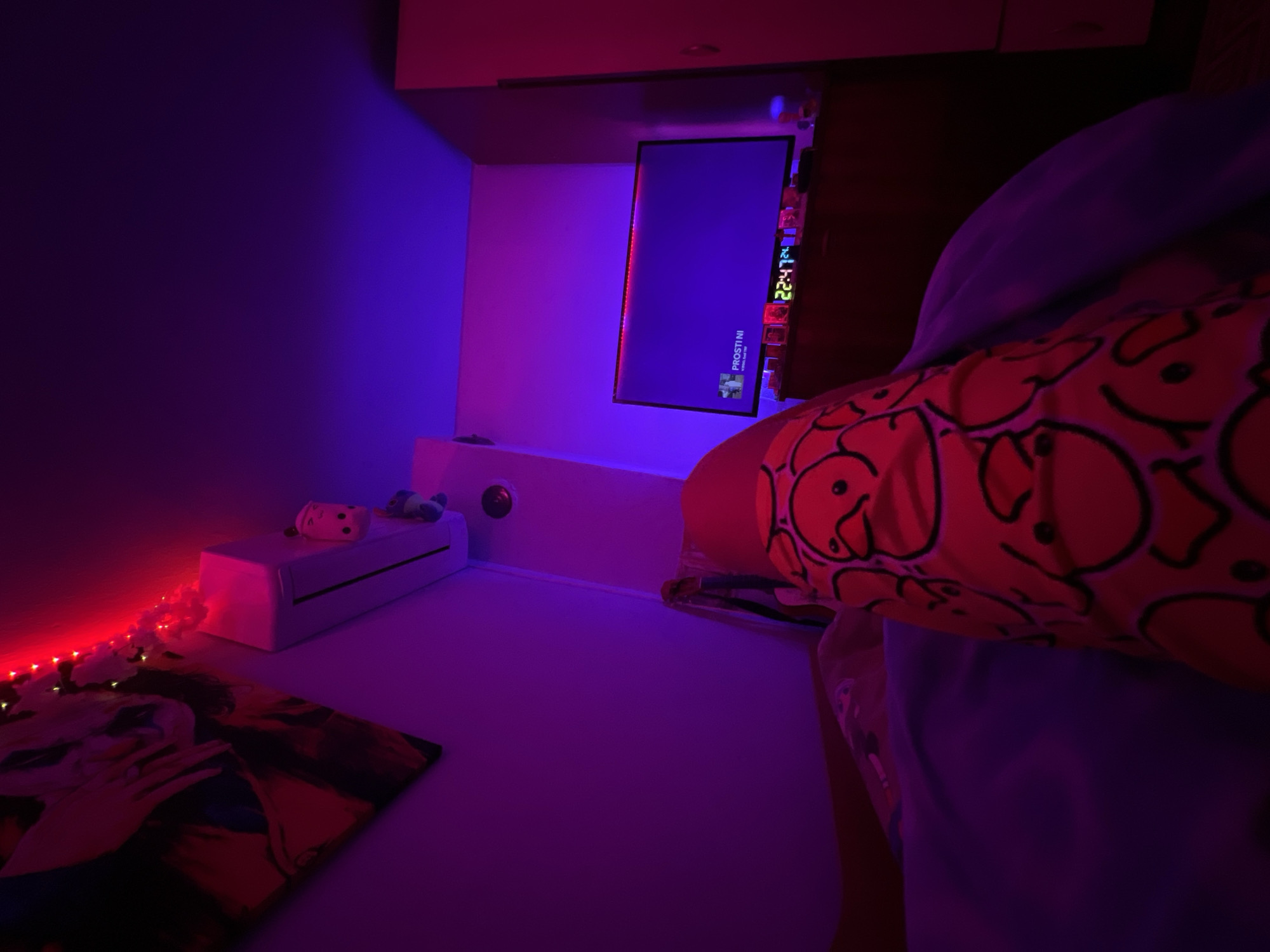 A bedroom interior, located in the city of Debelt, Bulgaria, is illuminated by prominent red and blue-purple LED ambient lighting. In the foreground, a person's legs are visible, partially covered by a light blue blanket. The visible leg is clothed in orange pajama bottoms featuring a repeating pattern of black-outlined ducks. On the upper left wall, a large, dark-toned framed artwork depicts a stylized figure. Below this artwork, a white wall-mounted air conditioning unit is adorned with a white and pink Funko Pop figure and a small grey plush toy. On the wall opposite the viewer, a flat-screen television displays a media interface with the text "PROSTINI" and "Yung N.". A red LED strip outlines the top and sides of the television screen. Below the television, on a dark wooden dresser, a digital clock clearly displays "22:47," surrounded by several small, indistinct objects that reflect the colored light. A light-colored, tall wardrobe or cabinet is partially visible on the far right side of the room.
