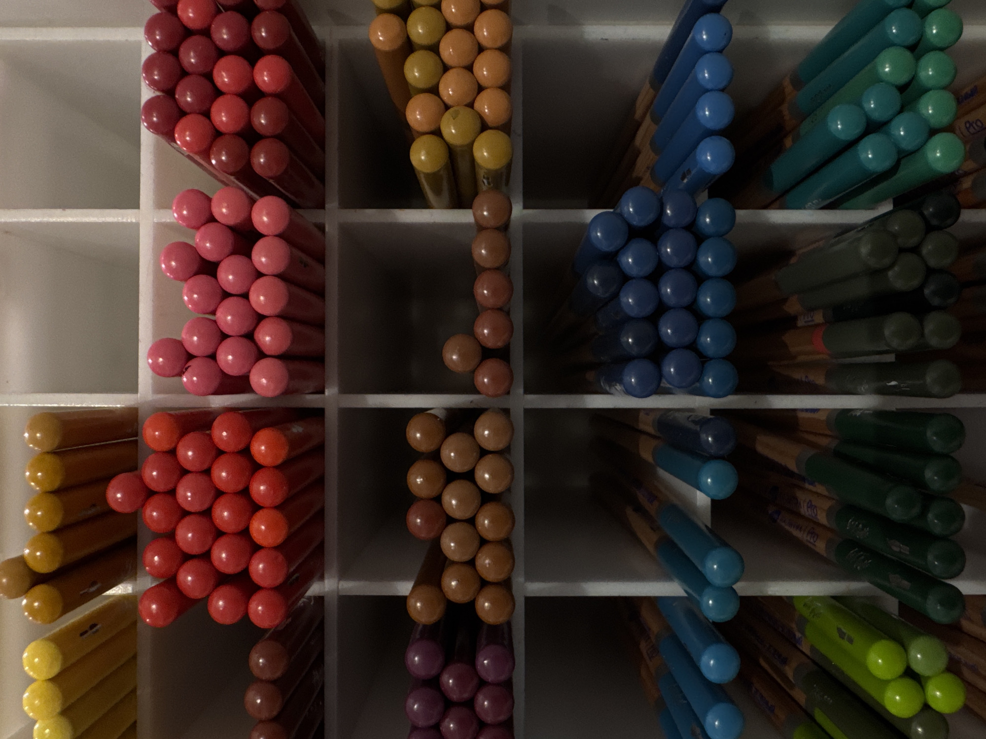 The image displays a close-up, top-down view of a white organizer filled with colored pencils. The pencils are arranged in neat rows and columns within the compartmentalized organizer. The colors visible include yellow, red, pink, purple, orange, brown, blue, and green. Some pencils have visible labels. The lighting creates shadows, highlighting the texture of the pencils and the organizer. No people are visible in the image. The scene is indoors, with a focus on the neatly stored art supplies. There is no information within the image to determine the location in Moscow, Russia.