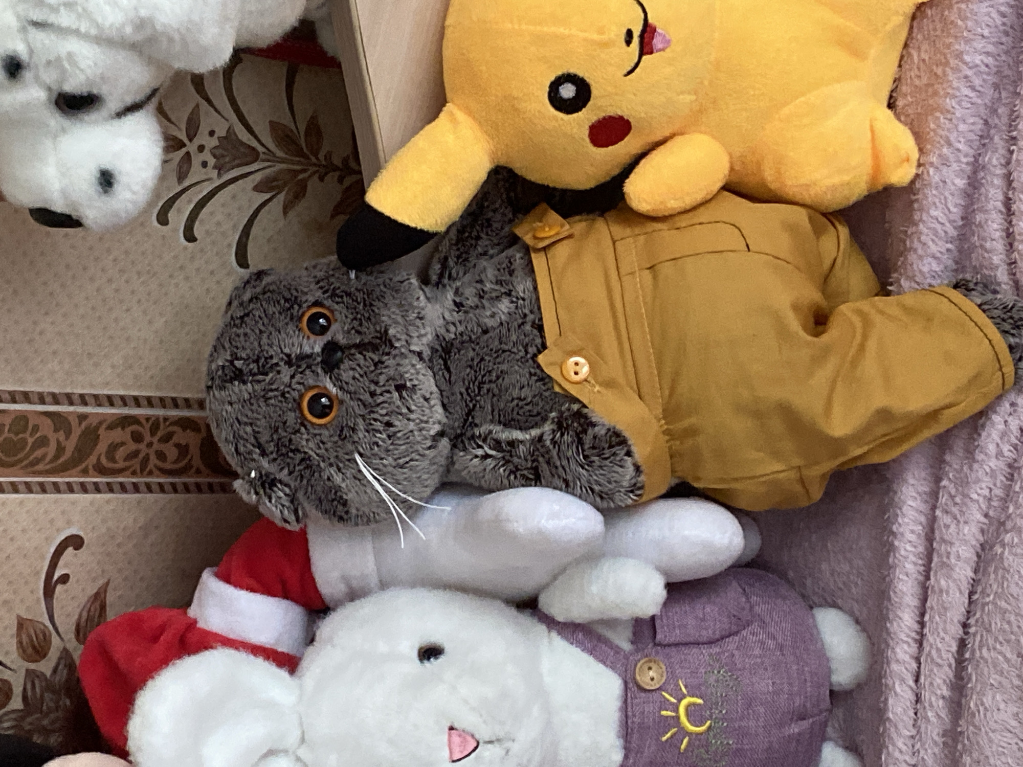 The image shows a collection of stuffed animals. In the foreground, from left to right, are a white rabbit wearing purple overalls with a sun emblem, a grey cat wearing yellow overalls, and a yellow Pikachu. Behind them, a plush figure in a Santa costume is partially visible, and a white plush dog is also present. The stuffed animals are arranged on a pink textured surface, possibly a blanket or carpet. The background features a patterned wallpaper. The scene appears to be indoors, within a residential setting. No specific location in Lomonosov, Russia is discernible from the image.