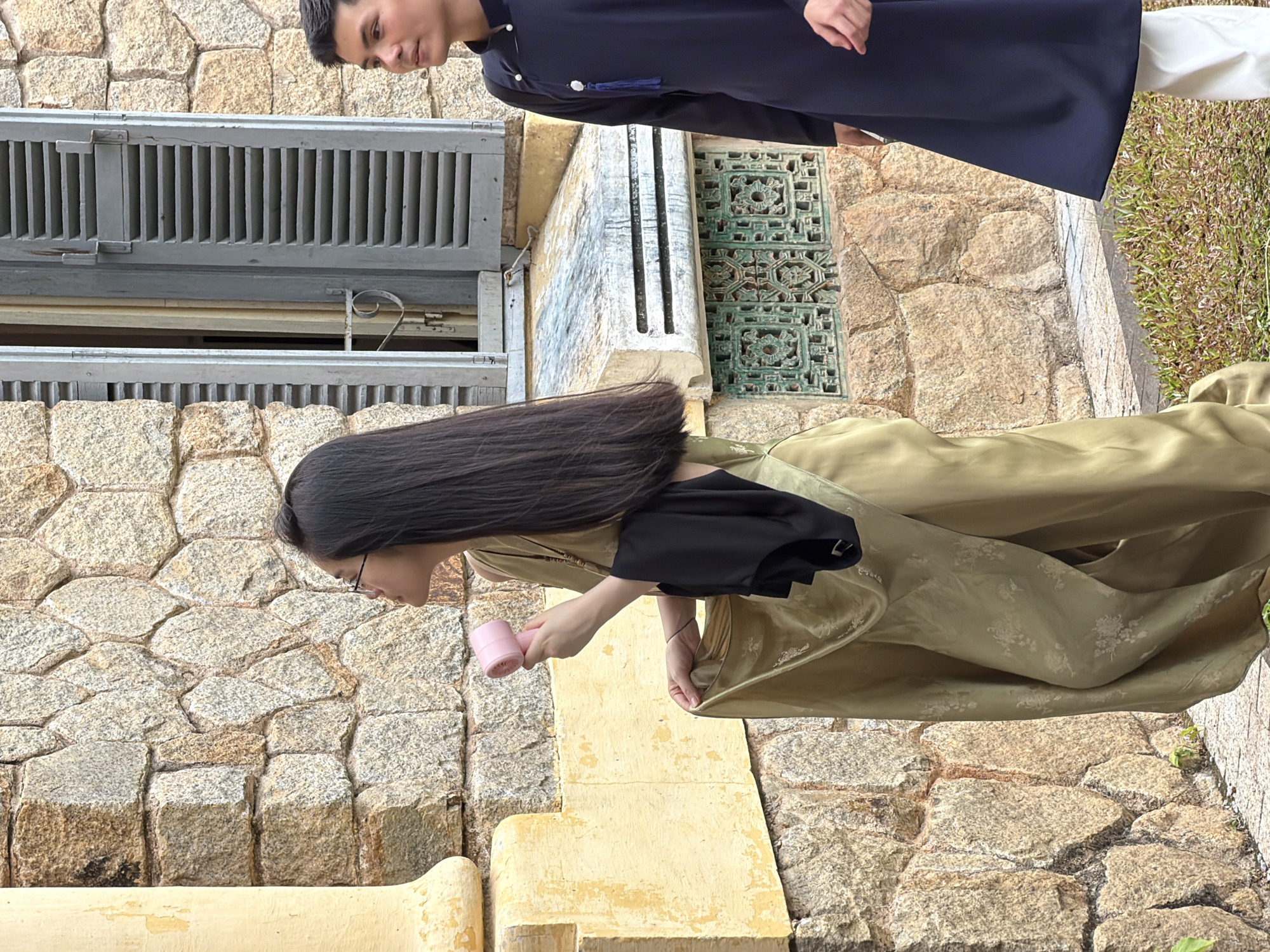 The image captures two individuals standing outdoors in front of a stone-built wall, possibly within the historic architecture of Thuận Hóa Ward, Vietnam. On the left, a person with long dark hair, wearing glasses, is holding a small pink fan. They are draped in a flowing, olive-green garment with subtle floral embroidery, and a black piece of fabric is tucked under their arm. To the right, a young man is dressed in a dark blue traditional Vietnamese tunic, with white trousers. He is looking to his left, towards the other individual. Behind them, a weathered window with grey shutters is visible against the stone wall, and below it, a decorative green grille is set into the stonework. The ground in front of them is a paved walkway with patches of green grass.