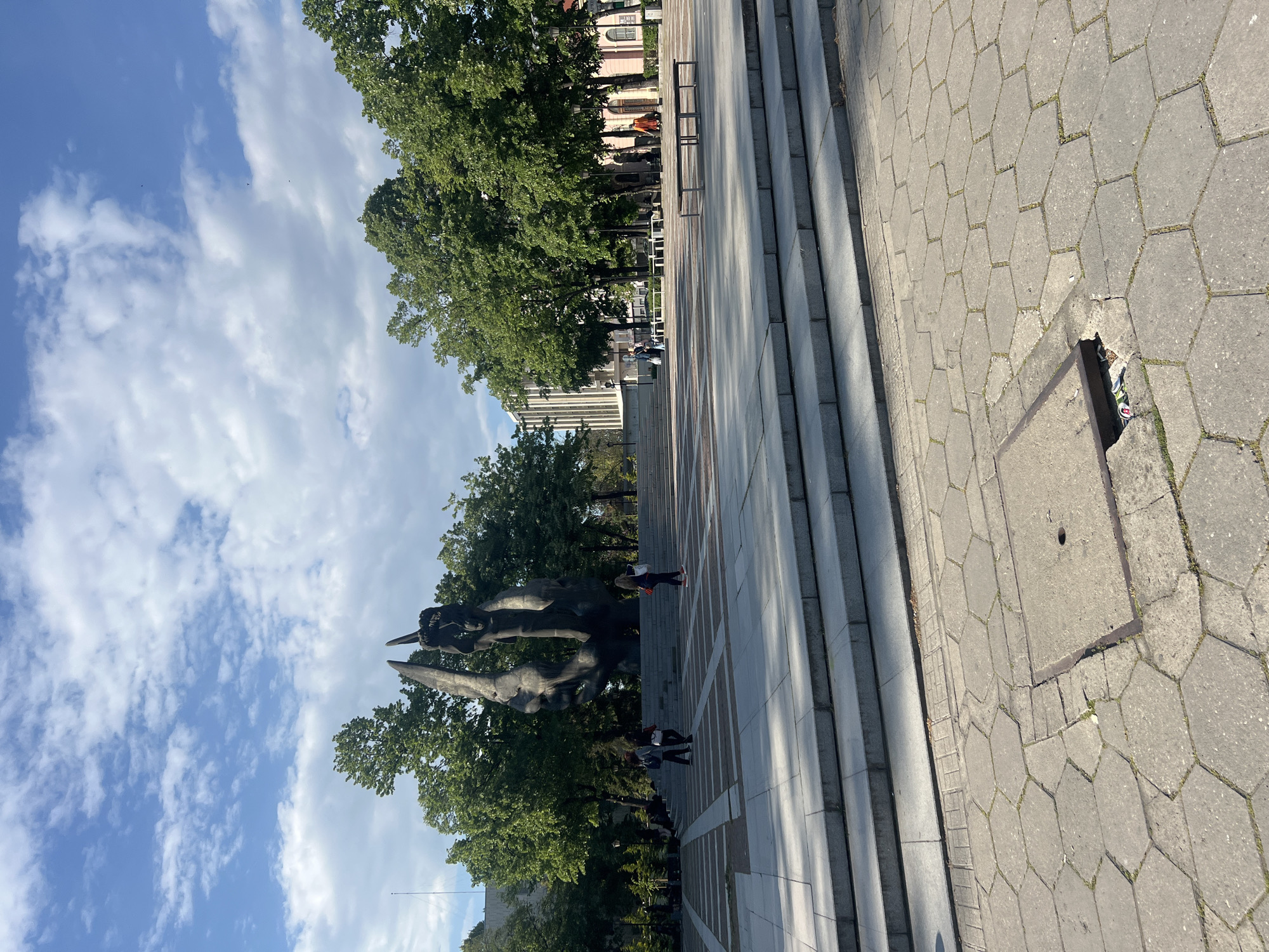 This image depicts an outdoor urban scene, likely a public square or park in Plovdiv, Bulgaria, under bright, clear conditions. The sky is a vibrant blue, dotted with scattered white, cumulus clouds, suggesting good weather during midday or early afternoon, as indicated by the short shadows.
The foreground and much of the right side are covered by a grey paved surface composed of hexagonal concrete tiles, with some weeds sprouting through the cracks. Along the left edge of this paved area, a wide, grey stone staircase ascends. Further right in the foreground, a broken section of the pavement edge reveals a small opening containing litter, including a crushed can. A square metal utility cover is also set into the hexagonal paving.
In the mid-ground, a large, dark-colored monument or sculpture stands prominently amidst numerous tall, green deciduous trees with full foliage, indicating a spring or summer season. The sculpture appears abstract or figurative, with a heroic scale, and is consistent with the "Monument to the Soviet Army" (Alyosha Monument) found in Plovdiv.
Several people are visible within the scene. One individual, wearing dark clothing and red shoes, is seen ascending the steps towards the sculpture. Another person stands near the base of the monument, and other smaller figures are present further in the background, walking among the trees and buildings.
Behind the line of trees, various buildings are visible in the distance, including older architectural styles with pitched roofs and more modern structures. Some metal street furniture, possibly benches or railings, is also present along a paved pathway in the background. The overall impression is one of a typical, calm day in a public gathering area. No discernible text is visible in the image.