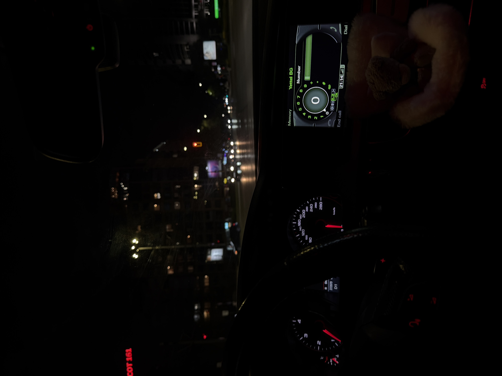 The image shows the interior of a car, at night, during a phone call. The infotainment system displays "Yettel BG Number" with a numeric keypad and the time is 21:14. The speedometer reads approximately 20 km/h, and the odometer shows 273979. A soft toy is placed to the right of the infotainment system. Through the windshield, city lights are visible, suggesting a nighttime urban environment. Red text visible on the left likely refers to something outside, such as street name COT 161. The image was likely taken in Sofia, Bulgaria, based on the "Yettel BG" displayed on the infotainment system, which is a Bulgarian mobile network operator.