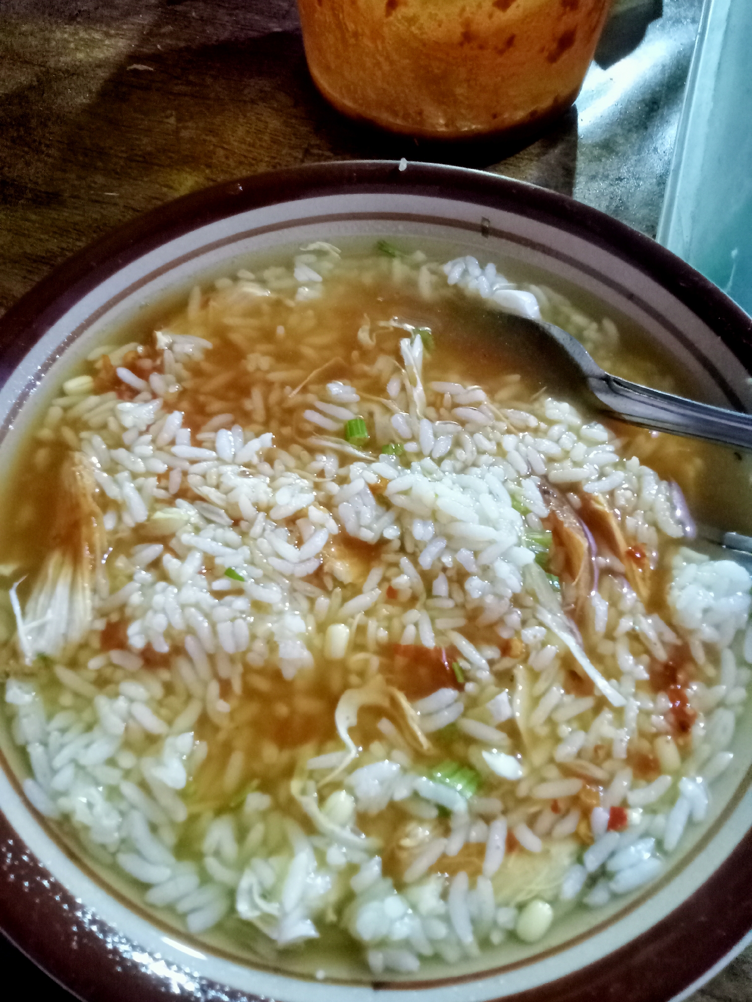 A close-up, high-angle shot shows a bowl of white rice with shredded chicken and green onions in a clear broth. The bowl has a brown and white rim. A silver spoon rests in the bowl, and a portion of an orange container is visible in the upper right corner. The background consists of a wooden surface. The scene is indoors, likely a casual dining setting in Wates, Indonesia.