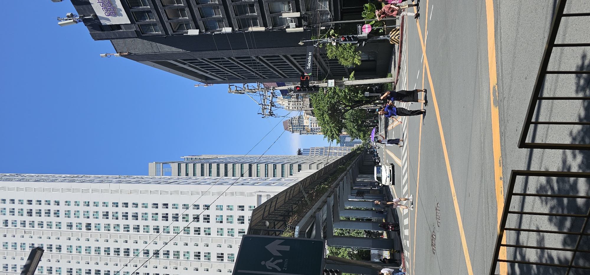 The image depicts a street intersection in Makati, Philippines, under a clear blue sky. Tall buildings dominate the cityscape. On the left, a modern, white high-rise building with a grid of windows stretches upwards. An elevated pedestrian walkway, supported by concrete pillars, is visible in the foreground, with a green directional sign indicating "2 min."
The intersection itself is paved with asphalt and marked with yellow lines. Several pedestrians are crossing the street. Two women in dark attire are walking across the intersection in the foreground. To the left, a man in a t-shirt and shorts is also crossing. Further back, a woman with a purple umbrella is walking, and other individuals and vehicles are present on the street.
Traffic lights and street signs are visible, including a sign for "Salcedo ST." The overall scene conveys a bustling urban environment in the Philippines.
