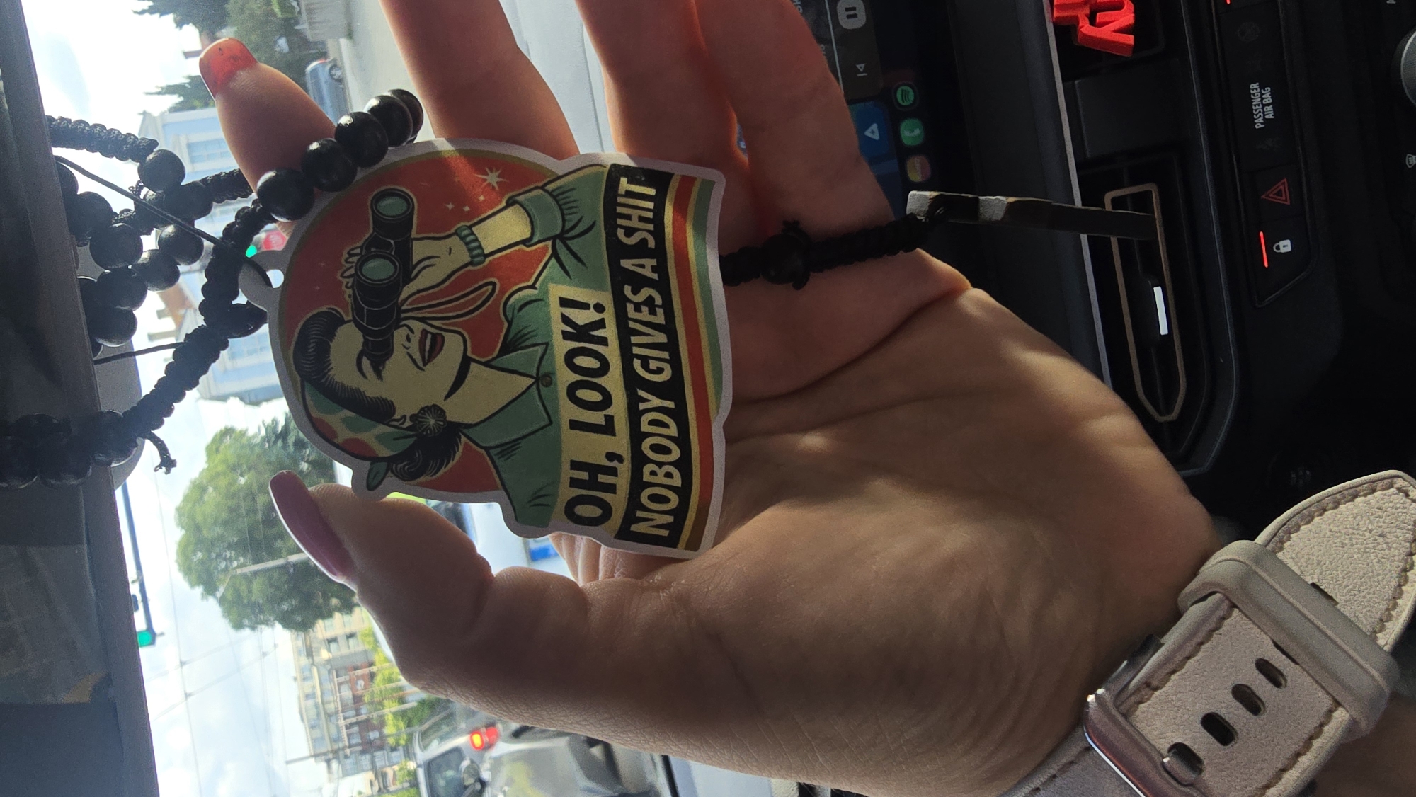 A hand holds a sticker with a woman holding binoculars that says, "OH, LOOK! NOBODY GIVES A SHIT," hanging from a beaded necklace in what appears to be a car. The location of the photo is Varna, Bulgaria. This is based on features of the surrounding architecture visible through the windshield of the car, which include distinctive, common urban construction, green trees, and sky. Additionally, the car's dashboard and interior are visible with the steering wheel and audio console. Also visible is a portion of a woman's watch on her wrist.