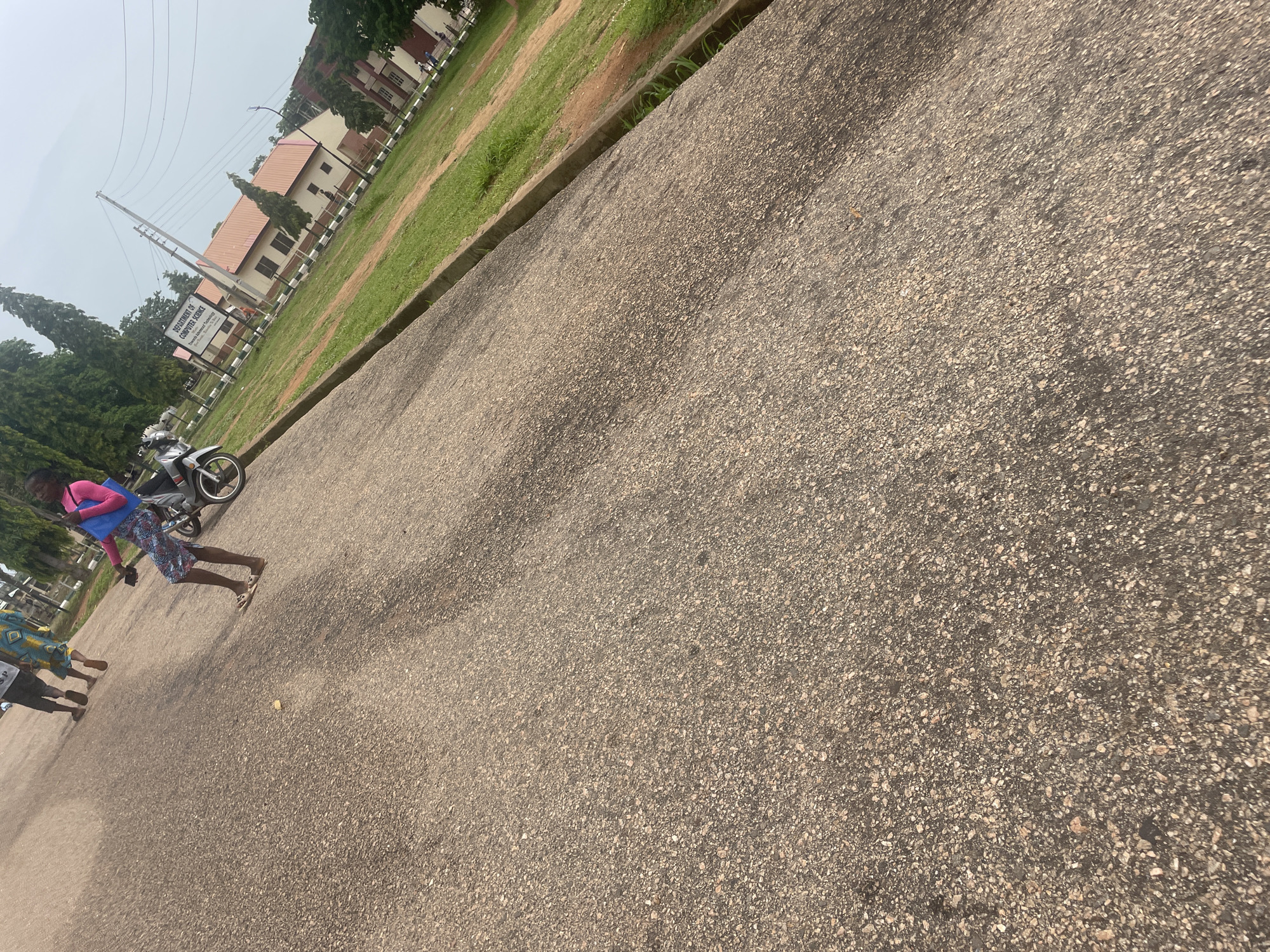 The image depicts an outdoor scene, likely on a paved road or walkway. The perspective is tilted, suggesting the photo was taken from a slightly elevated or angled position. In the foreground, the textured surface of the pavement dominates the frame.
Further back, several figures are visible. One person, dressed in a bright pink top and a patterned skirt, walks alongside a motorcycle. This individual is carrying a blue bag. Another person, partially visible in the lower left, is also walking. A signpost is also visible in the distance, which reads: "DEPARTMENT OF COMPUTER SCIENCE" and below that, "Faculty of Science". Trees and buildings line the background, suggesting an institutional or campus setting. The sky appears overcast, hinting at a cloudy or hazy day.
The overall impression is of a casual moment captured outdoors, possibly on a university campus or in a similar environment in Lokoja, Nigeria. The lighting suggests it is daytime. There are no strong indicators of a specific event, but rather a snapshot of everyday activity.
