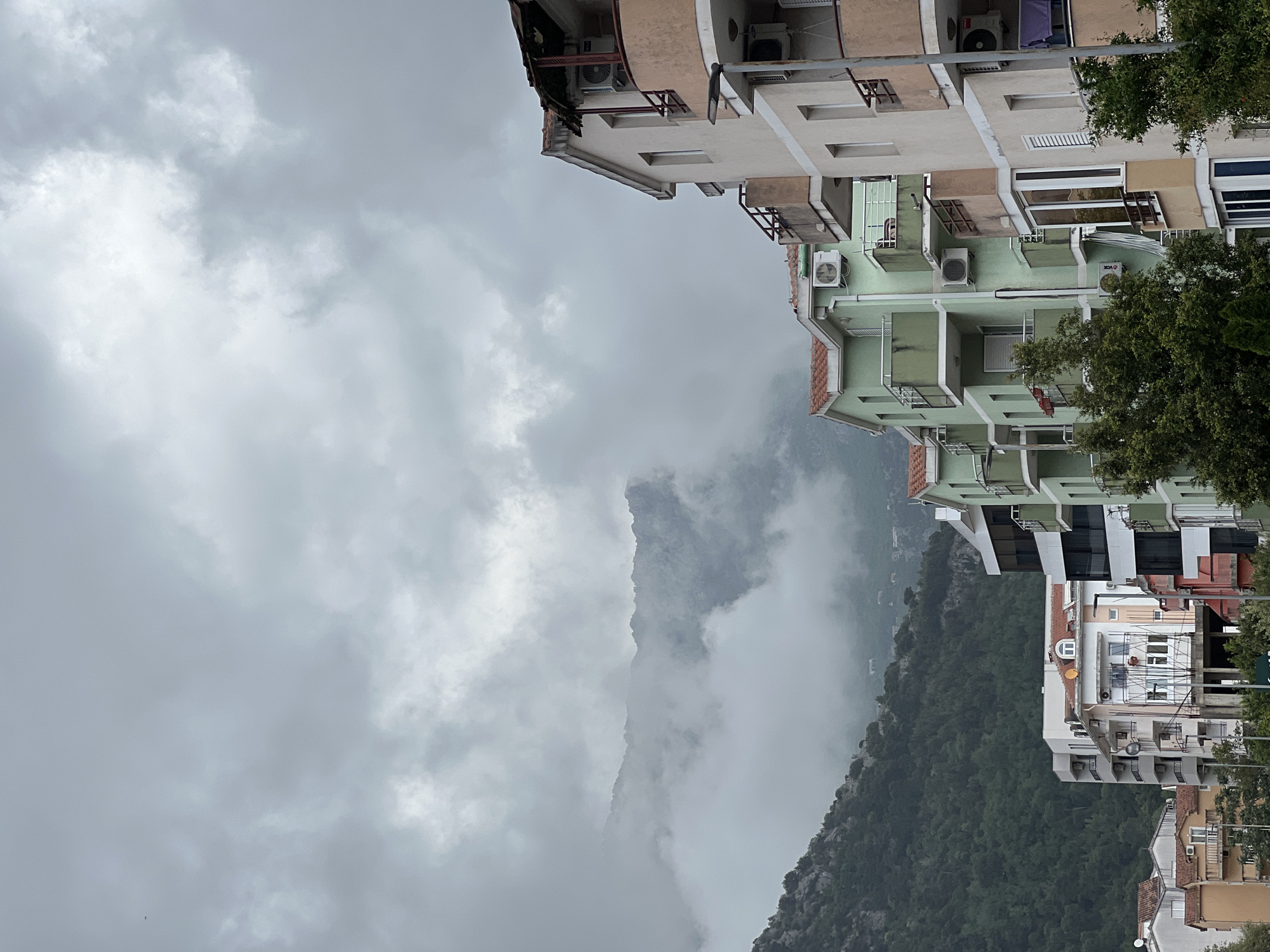 The image shows a view in Budva, Montenegro, on a cloudy day. A rugged mountain range is partially obscured by mist. In the foreground are several multi-story apartment buildings, including light brown, green, and white ones, arranged vertically. The buildings have balconies and air conditioning units. Trees are visible among the buildings. The sky is overcast with dense, grey clouds. The picture is taken from a high angle, looking down and out towards the mountains.
