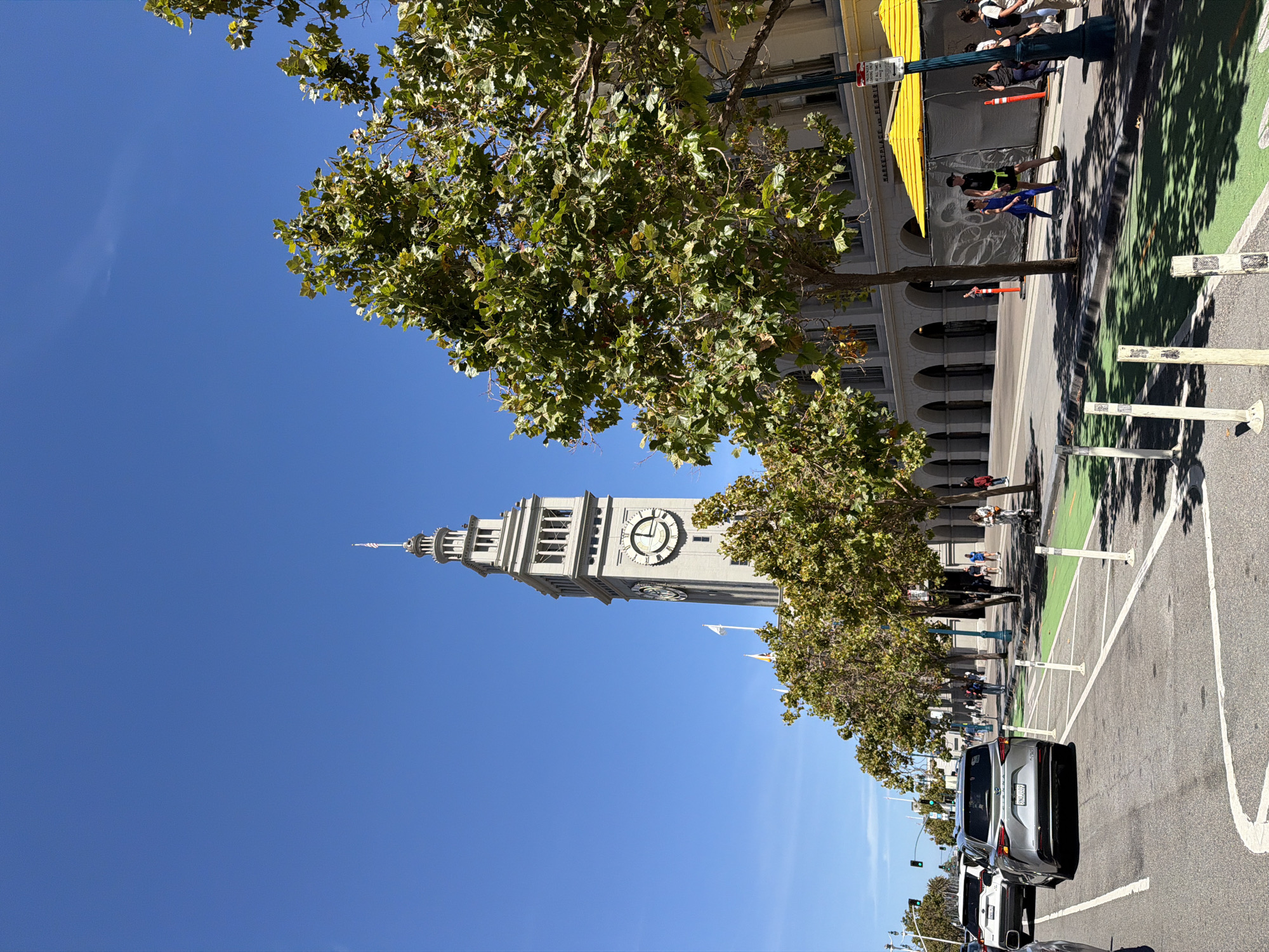 The image shows a view of the San Francisco Ferry Building, a landmark located at the foot of Market Street in San Francisco, California. The Ferry Building is a distinctive Beaux-Arts style building with a prominent clock tower, visible in the background under a clear blue sky. In the foreground, a tree-lined street is depicted with cars driving on the right side of the road. A dedicated bike lane, painted bright green, is present on the right side of the road, separated from the car traffic by white bollards. Pedestrians are seen walking on the sidewalk in front of the Ferry Building, some under yellow awnings. The scene captures a sunny day in the city.