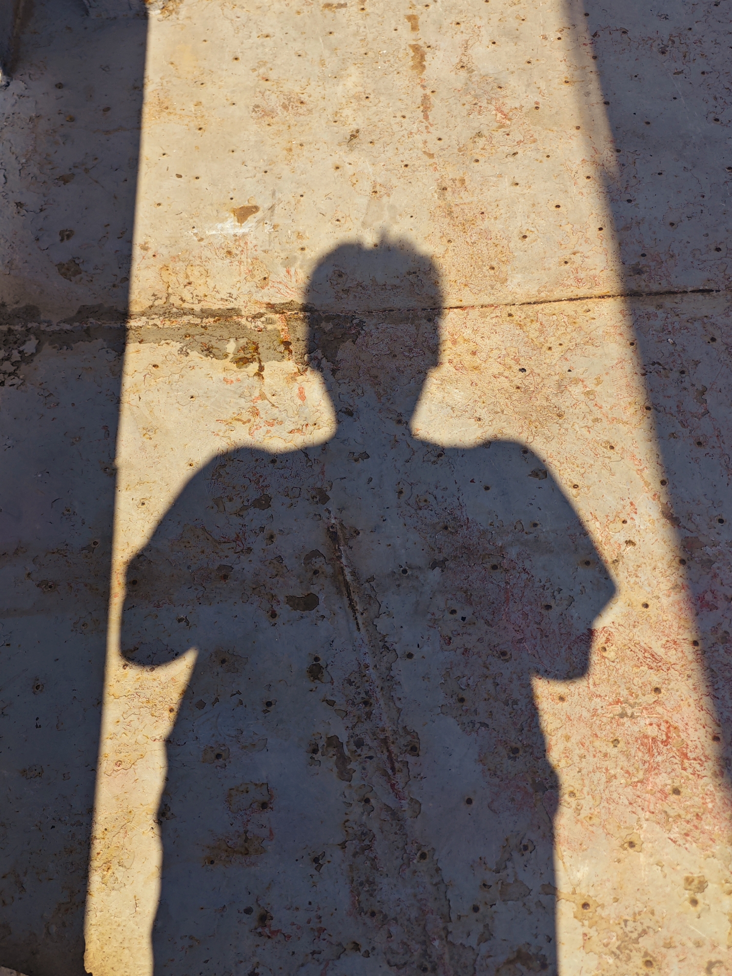 A shadow of a person with arms crossed is cast on a rusted, textured surface. The surface appears to be metal, possibly weathered or industrial, with patches of rust and peeling paint. A bright light source, likely the sun, creates a sharp, diagonal shadow that dominates the lower left portion of the image, with a second, fainter shadow to the right. The environment is outdoors, in an urban setting, with the distinct texture of the ground suggesting it might be a weathered metal platform or walkway.
