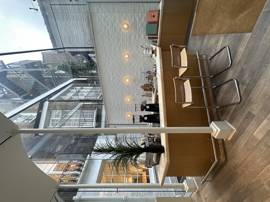 The image displays the interior of a coffee bar, identified as The Gentlemen's Baristas on Lamb's Conduit Street, Bloomsbury, London, United Kingdom. The space is enclosed by a large glass ceiling and wall structure, looking into an urban courtyard.
In the foreground, dark wooden floorboards are visible. Two chrome-framed bar stools with woven cane seats face a light-colored wooden coffee counter with a beige marble top. On the counter, from left to right, are a black espresso machine, a tall black vase containing green faux foliage, two black conical coffee grinders, various metal barista tools, and a sink. A transparent cloche covers items on the right side of the counter, adjacent to a lower wooden cabinet section displaying colorful books.
The wall behind the counter is constructed from white-painted brick, featuring five evenly spaced, circular, illuminated wall sconces. White, fluted structural columns support the glass enclosure.
Through the glass, the exterior courtyard reveals multi-story brick buildings with numerous windows, balconies with black iron railings, and an external black metal spiral fire escape on the right side. Green foliage is visible near the fire escape. The sky overhead is overcast. No people are present in the scene. The area is clean and arranged for commercial coffee preparation.
