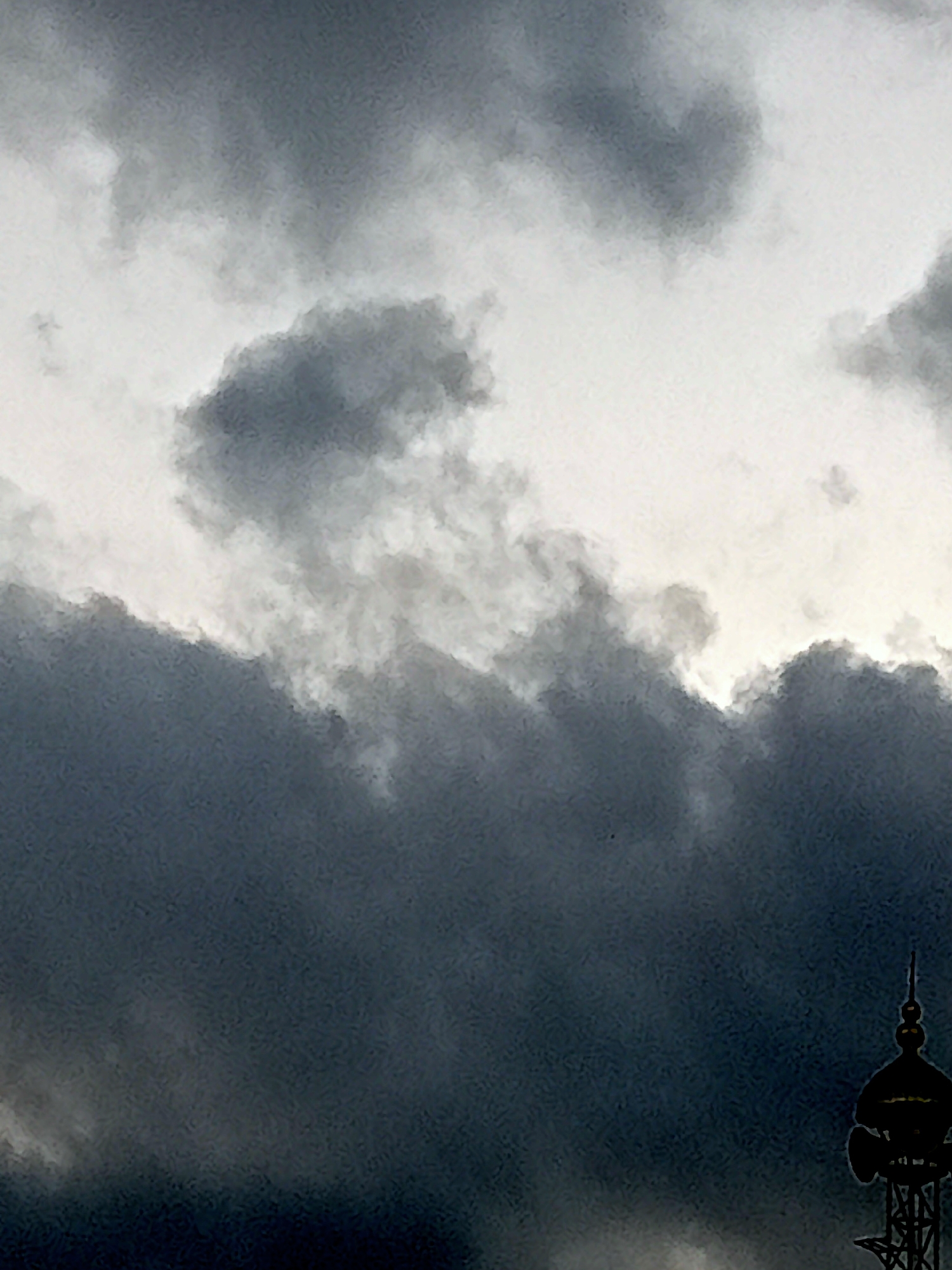 The image depicts a dark, overcast sky with heavy, textured clouds. In the lower right corner, the silhouetted top of a tower is visible against the cloudy backdrop. The tower features a domed structure with a pointed spire and multiple levels with visible framework and what appear to be speakers or lights. The overall impression is of a dramatic, possibly stormy, sky over an urban environment. The location in Simo, Indonesia is not explicitly visible or derivable from the image itself.