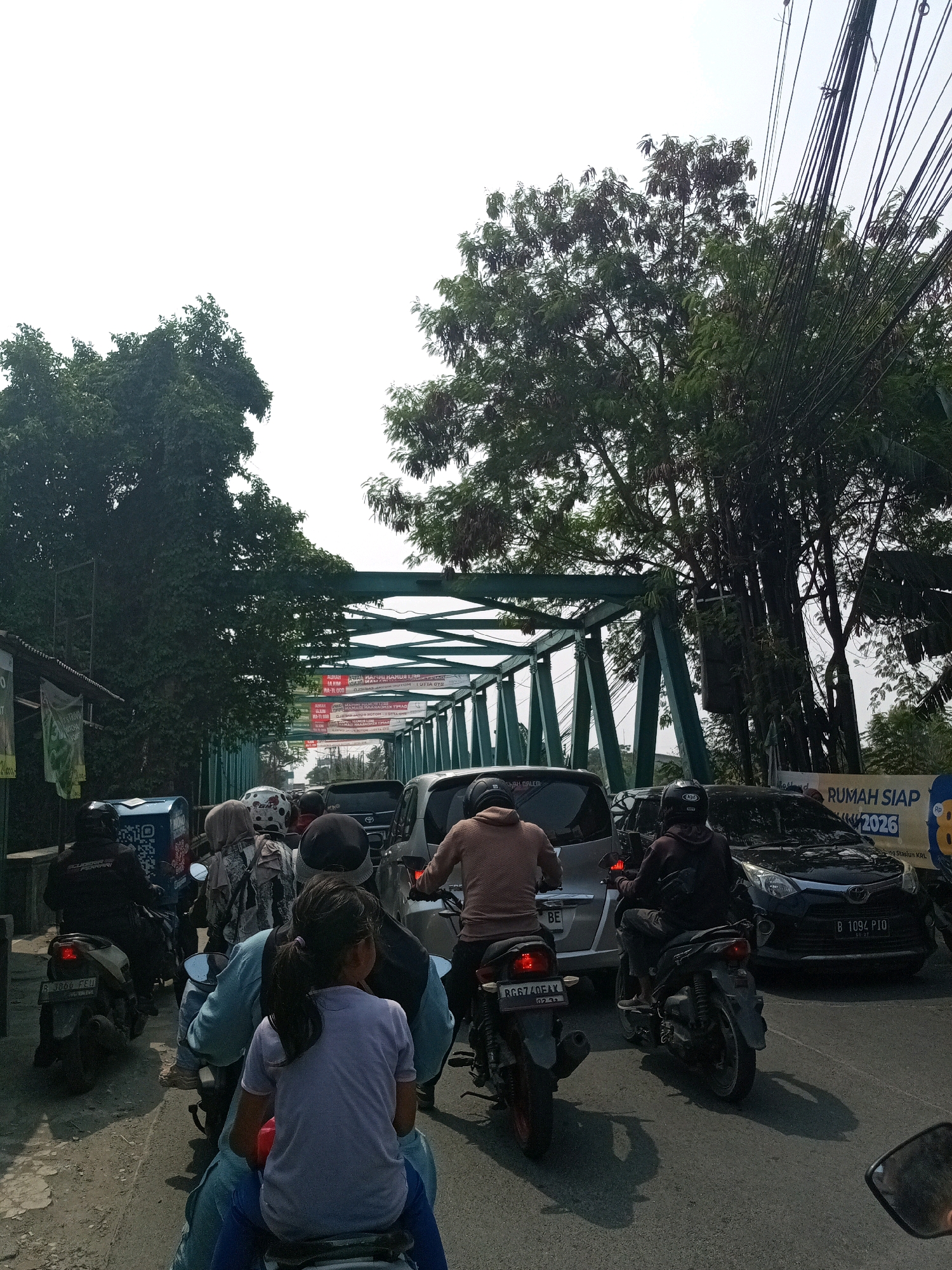 In Karangsatria, Indonesia, a traffic jam unfolds beneath a green metal truss bridge. On the road, multiple motorcycles and cars are stationary. In the foreground, a child wearing a purple t-shirt and blue pants sits on the back of a motorcycle, facing away from the camera. The motorcycle is ridden by an individual in a light blue shirt. Another motorcycle to the left is ridden by a person in a black jacket. Behind them, a woman in a patterned headscarf is seated on a motorcycle. To the right, a car with license plate B 1094 PIO is visible, and further back, another car with a license plate that appears to read B 3366 FFI is also stopped. Overhead, a dense network of power lines crisscrosses the sky, with lush green trees framing the scene. Billboards with Indonesian text are present on the side of the road and on the bridge structure. The lighting suggests it is daytime, with sunlight casting shadows on the asphalt.