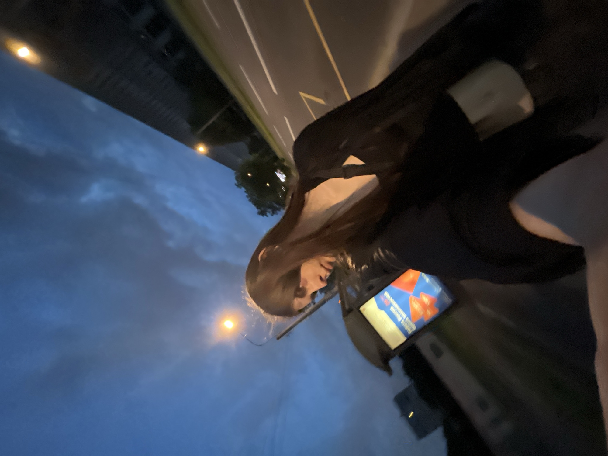 The image depicts a nighttime urban scene in Saint Petersburg, Russia, captured from a low, slightly tilted angle, suggesting a selfie perspective. In the mid-ground, a female individual with long, dark brown hair is visible, looking downwards and to her left. She is dressed in dark clothing, possibly a black jacket or blazer over a dark top, with a light-colored bag partially visible on her right side. Her right arm and hand, likely holding the camera, are present in the lower foreground.
To the left of the individual, a public transport stop shelter features an illuminated advertisement panel. The advertisement is in Russian, displaying "Почта России" (Russian Post) and "Отправляй посылки" (Send parcels), accompanied by red, stylized graphic elements. Above the bus stop, a streetlight emits a warm, yellow-orange light, illuminating the immediate area. Another similar streetlight is visible further down the road on the right.
The background on the right reveals a multi-lane asphalt road marked with distinct white dashed lines and a continuous yellow line, along with a yellow chevron-shaped marking. In the upper right background, a large, dark, elongated building or elevated structure runs parallel to the road. The sky is dark blue with visible cloud formations, indicative of dusk or night. Dark tree foliage is present in the mid-distance to the left. The presence of the "Почта России" advertisement confirms the setting within Russia, specifically Saint Petersburg as per the prompt's instruction.