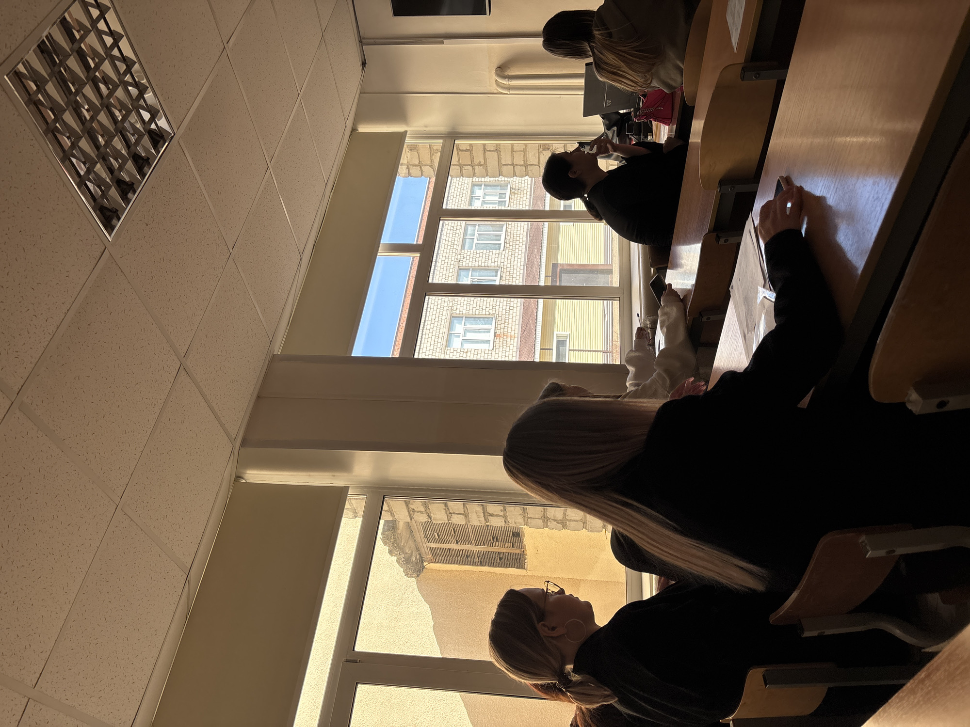The image shows a group of students in a classroom setting, likely a university lecture hall. The classroom is illuminated by natural light streaming through windows. Outside, there is a view of buildings, including a multi-story building made of brick, along with other structures, suggesting an urban environment.
The classroom has rows of desks, some occupied by students who appear to be engaged in different activities. At least one person is using a mobile phone.
Given the location, the image suggests a scene taking place in an educational institution within the city of Chaikovsky, Russia.