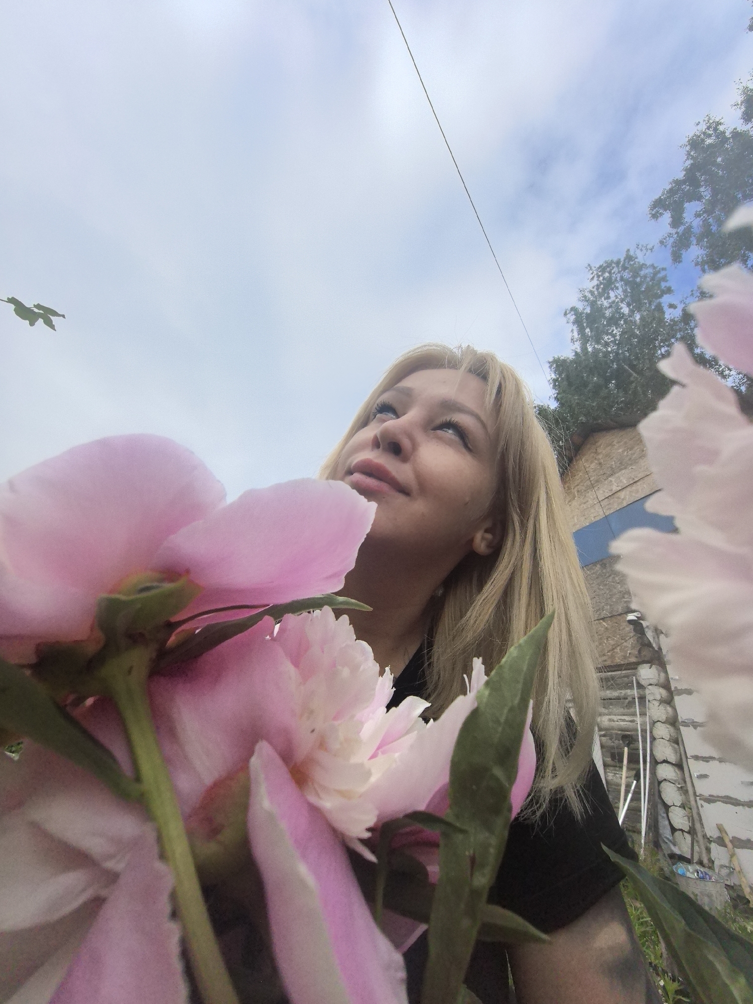 A low-angle, medium shot captures a young woman with blonde hair looking upwards with a slight smile, framed by large pink peonies in the foreground. The woman is wearing a black shirt and has tattoos on her arm. Behind her, a rustic wooden structure and lush green trees are visible under a cloudy sky with a thin wire stretched across it. The scene appears to be outdoors, possibly in a rural or suburban area. While the image does not explicitly state the location, the query specifies Noviy, Russia, placing the context of the scene within that locale.