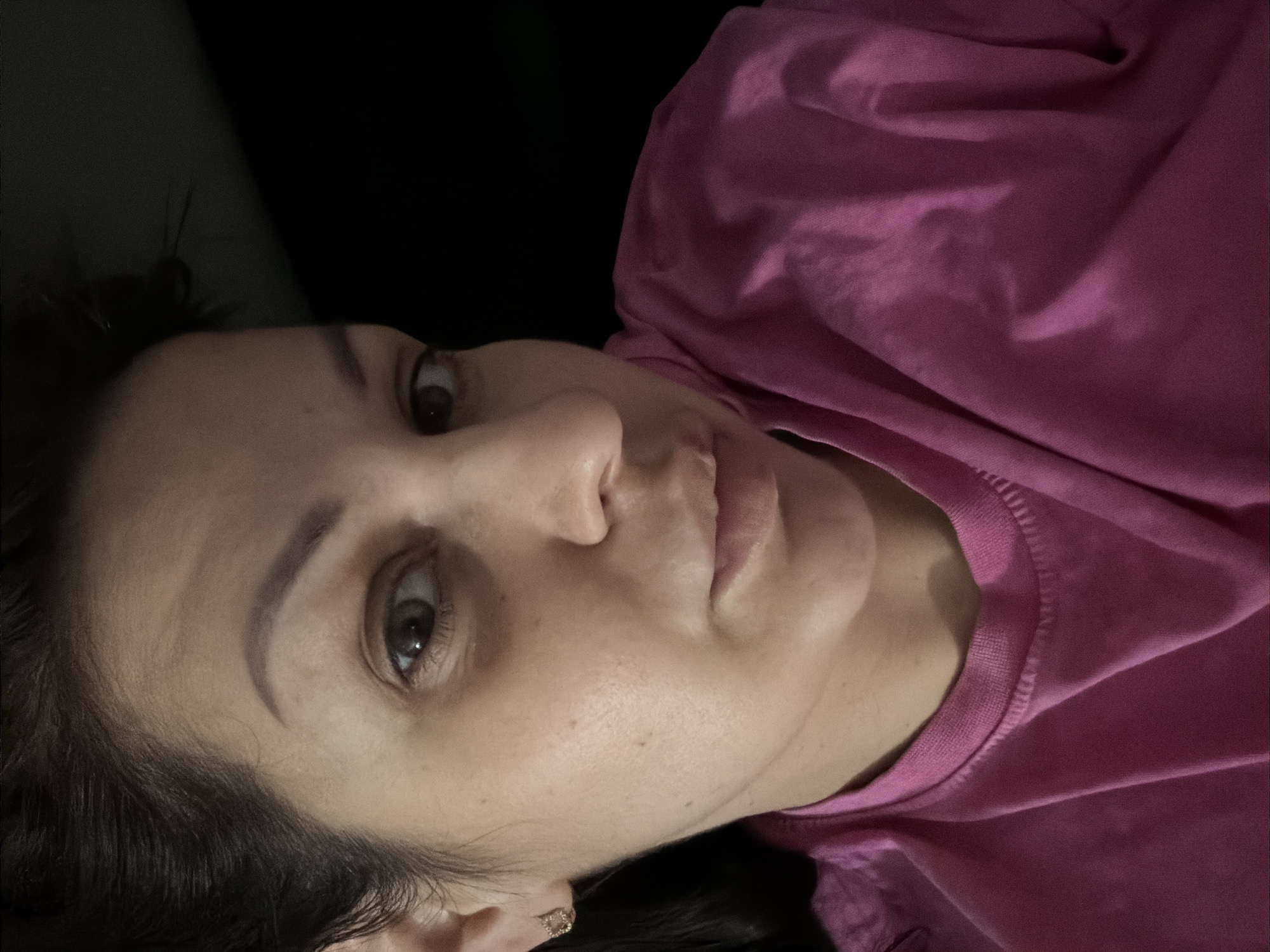 A close-up, low-angle selfie shows a woman with dark hair and brown eyes wearing a pink t-shirt. She is looking directly at the camera with her lips slightly parted. The background is dark and out of focus. There is no indication of the city of Sochi, Russia, or any specific location within a city.