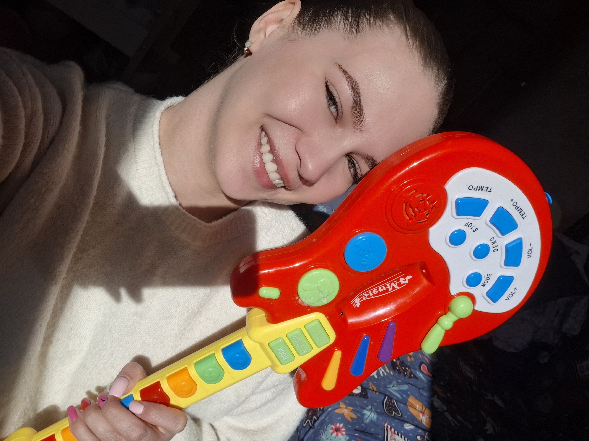 A young woman with fair skin, dark hair pulled back, and a wide, joyful smile is captured in a close-up image, likely indoors in Chaikovsky, Russia. Her head is tilted to the left, resting against a brightly colored, red and yellow plastic toy guitar that she holds with her right hand.
The woman wears an off-white, textured knit sweater. Her fingernails are painted in alternating shades of pink. A small earring is visible in her right ear. A strong, directional light source, likely natural light, illuminates her face and the left side of her body, casting distinct shadows across her sweater and forehead.
The toy guitar features a red body and a yellow neck adorned with various colorful buttons and controls. On the red body, a white panel contains blue buttons labeled "TEMPO+", "TEMPO-", "STOP", "DEMO", "MODE", "VOL+", and "VOL-". A "♪ Music" logo is visible near the neck joint. The yellow neck has three green rectangular buttons labeled "ROCK", "HIP HOP", and "JAZZ", along with other colorful round and oval buttons (blue, green, light blue, purple, yellow) resembling musical notes or frets. A small, light-green microphone-like attachment extends from the red body. The background is dark, with a patterned blue fabric, possibly a blanket, visible to the right. The woman's expression conveys happiness and contentment as she interacts with the toy.