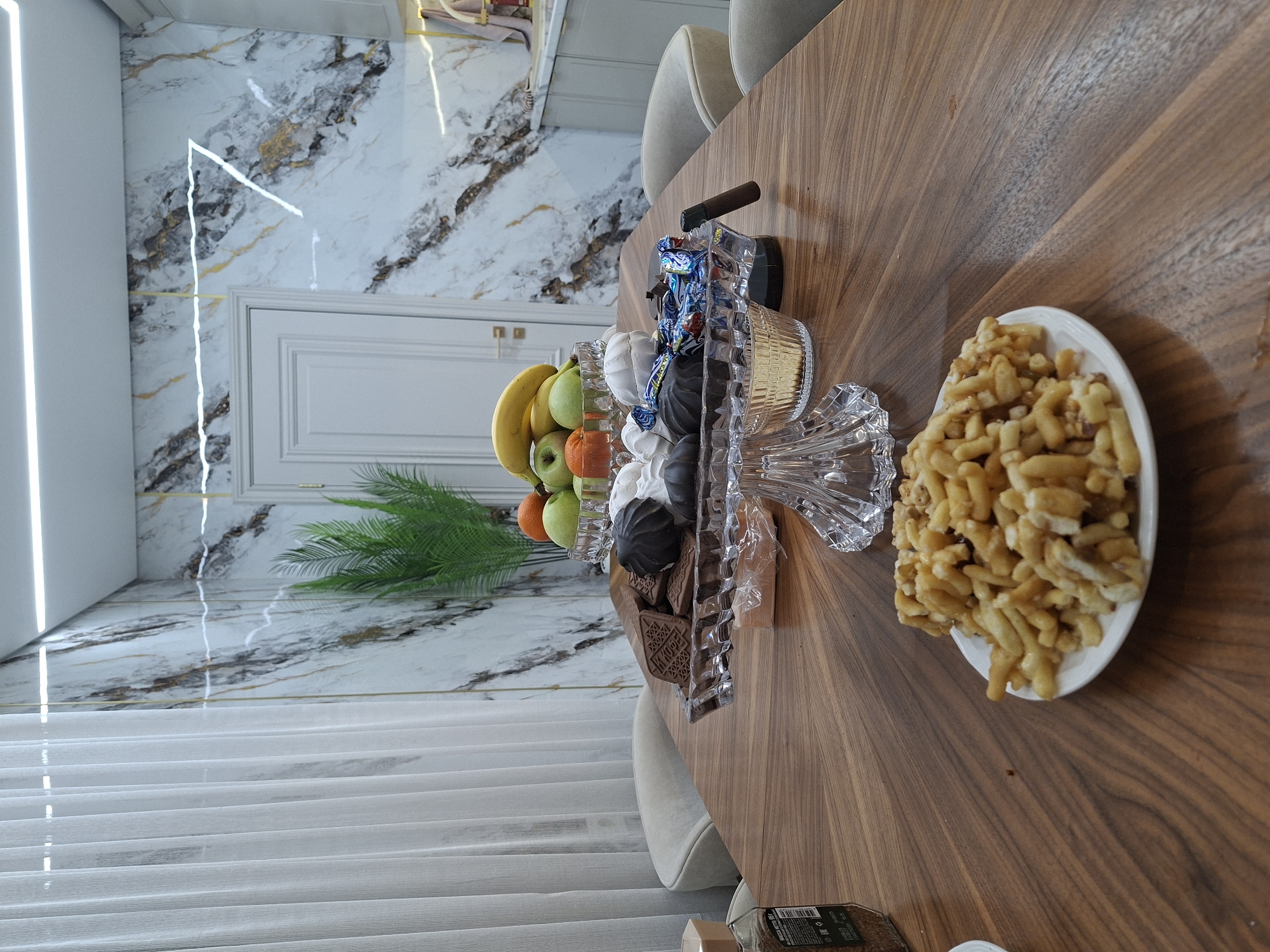 Here is a detailed description of the image:
The image depicts a dining table laden with food and decorative items. On the table, there's a crystal bowl filled with fruits, candies, and chocolates, as well as a plate of fried dough pastries. A cigar rests on top of the bowl, and a green plant is situated nearby. The table appears to be made of wood with a visible grain.
The background consists of a marble wall with light beige accents and a partially visible white door. Sheer curtains are hanging in front of a window, allowing soft light to filter into the room. The overall setting suggests an indoor dining area with a luxurious aesthetic. The location is specified as Unknown, Unknown.
