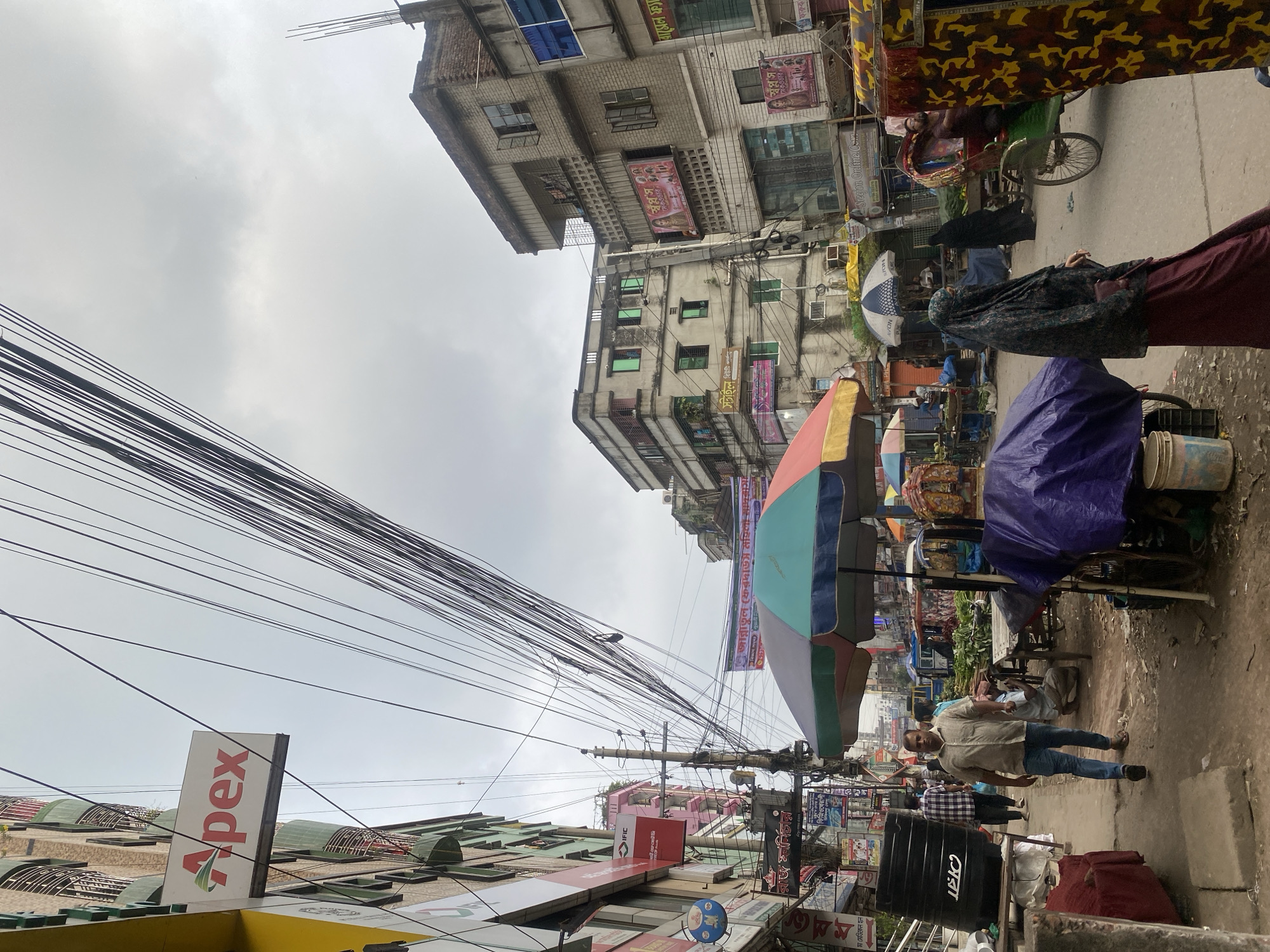 This is an outdoor, eye-level, full-shot photograph of a bustling street scene in Dhaka, Bangladesh, likely a market area. The sky is overcast, creating a diffused light. On the left side of the image, a multi-story building features various signs, including an "Apex" logo. Buildings on both sides of the street are densely packed, with numerous electrical wires crisscrossing overhead, a common sight in Dhaka.
In the foreground, a man in a light-colored shirt and blue jeans walks towards the viewer. To his right, a woman wearing a dark hijab and a long, deep red skirt stands facing away from the viewer. Behind her, a colorful rickshaw is being pushed by an individual. Further down the street, market stalls are visible with umbrellas providing shade over goods, likely produce. Several people are walking and interacting within the market. The ground is paved, and there are various objects scattered around, including a large black plastic tank on the left, and buckets near the market stalls. The overall impression is of a vibrant and active urban environment, characteristic of Dhaka's busy commercial districts.