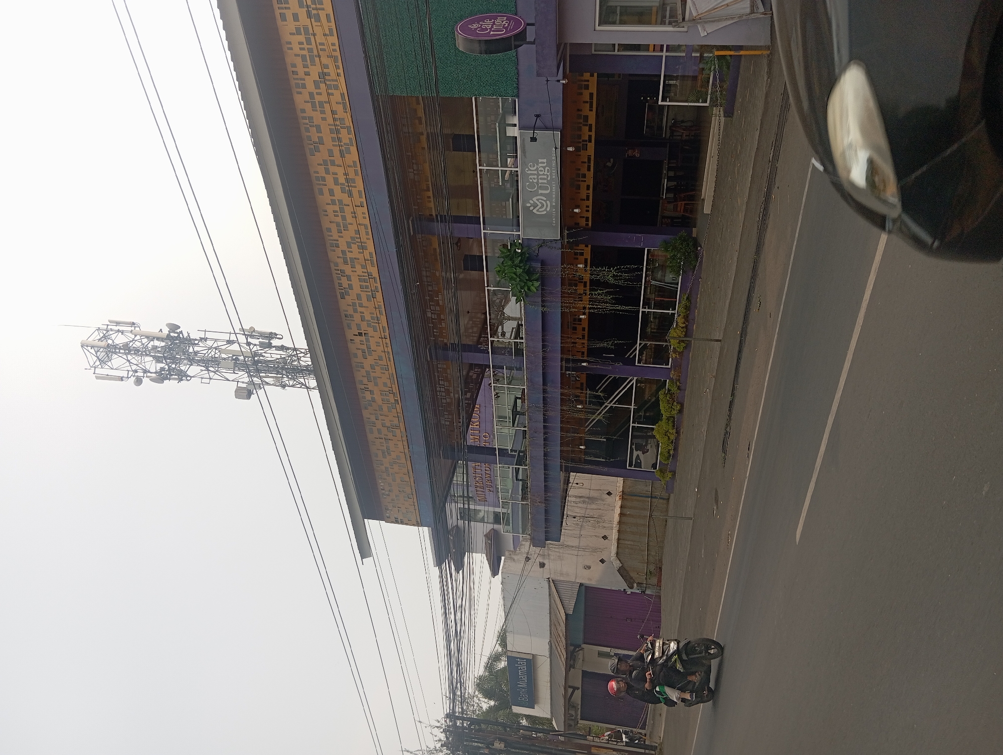 In Sumampir, Indonesia, a street scene captures a multi-story building with a telecommunications tower atop it. The building features a distinct yellow patterned facade on its upper level and purple accents on its lower levels. A sign for "Cafe Ungu" is prominently displayed, suggesting it is a cafe, minimarket, and meeting room. To the left, a sign for "Bank Muamalat" indicates the presence of a bank. A motorcycle carrying two individuals, one wearing a red helmet, travels along the asphalt road in the foreground. The sky is overcast, contributing to a hazy atmosphere.