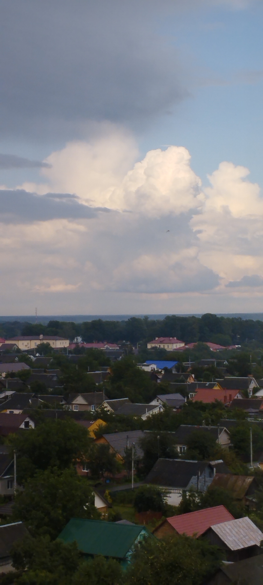 An aerial, vertical shot captures a dense residential area, likely Skidzieĺ, Belarus, under a dramatic sky. The upper portion of the image is dominated by a turbulent cloud formation, with dark grey clouds on the left transitioning to bright white, puffy cumulus clouds illuminated by sunlight on the right, against a pale blue sky. A very small dark speck, possibly a distant bird or drone, is visible within the clouds.
The lower half of the image depicts a sprawling, low-rise urban landscape. Numerous houses with varied roof colors and styles are nestled amongst dense green trees. The rooftops range from dark grey, brown, and red tiles to a distinctive bright green roof in the foreground. Several larger, institutional-looking buildings with yellow and red facades are visible in the mid-ground, interspersed with the smaller residential structures. A bright blue roof stands out on one of the mid-ground buildings. The overall impression is of a densely populated, established neighborhood with mature vegetation. The lighting suggests it is either early morning or late afternoon.