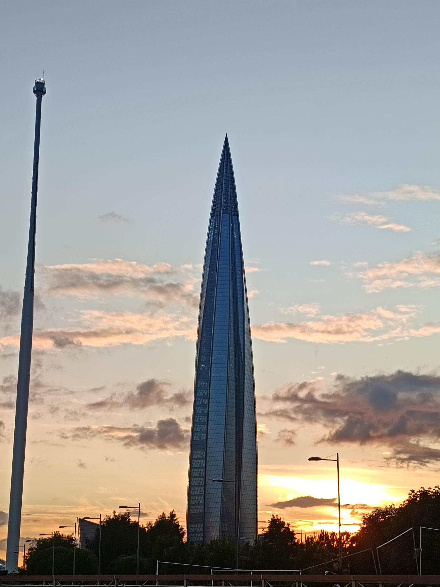 The image captures a sunset scene in Saint Petersburg, Russia, featuring the distinctive Lakhta Center skyscraper. The tall, slender, glass-covered skyscraper dominates the right side of the frame, its pointed apex reaching towards the sky. To the left, a tall, slender tower with a small structure at its top stands prominently. The sky is a canvas of soft blues, pinks, and oranges, indicative of sunset, with scattered clouds tinged with warm hues. Silhouetted against the horizon are trees and streetlights, suggesting a city park or embankment. A fence line runs across the foreground, with indistinct figures visible near it, possibly pedestrians or onlookers enjoying the evening view. The overall atmosphere is serene and picturesque, highlighting a modern architectural marvel against a natural sunset backdrop.
