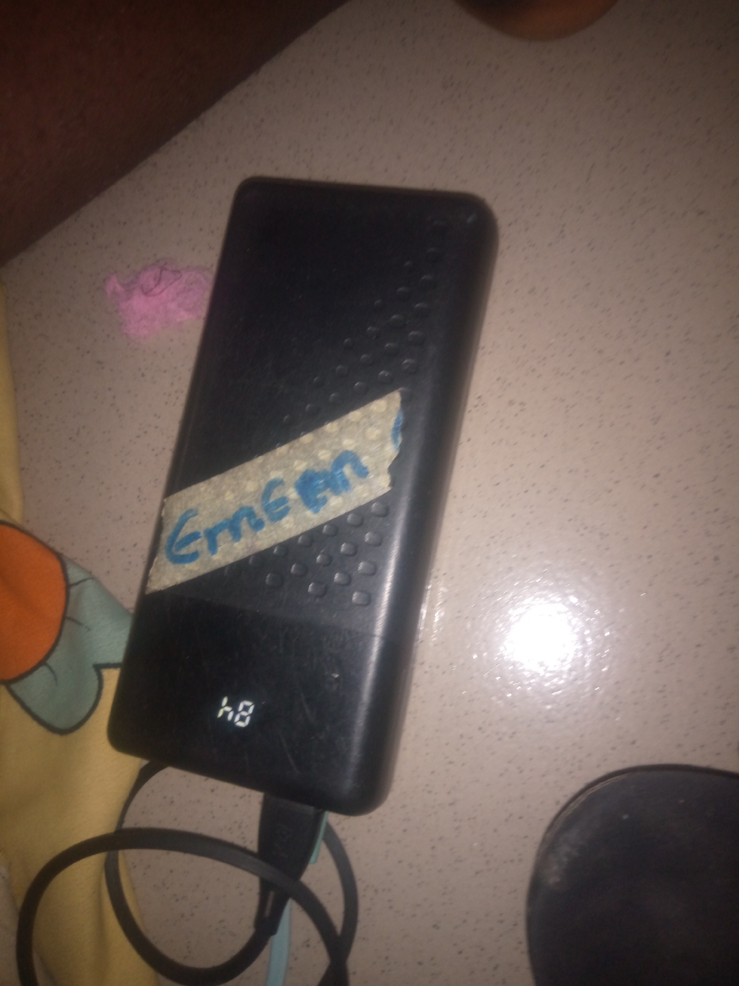 A black portable charger with the word "Emeen" written on it in blue marker on a piece of taped-on paper is resting on a speckled beige countertop. The charger has a digital display showing "48". A black charging cable is connected to the charger and coiled around it. To the left of the charger, a yellow and orange patterned cloth is visible. In the background, a small piece of pink material is on the countertop.