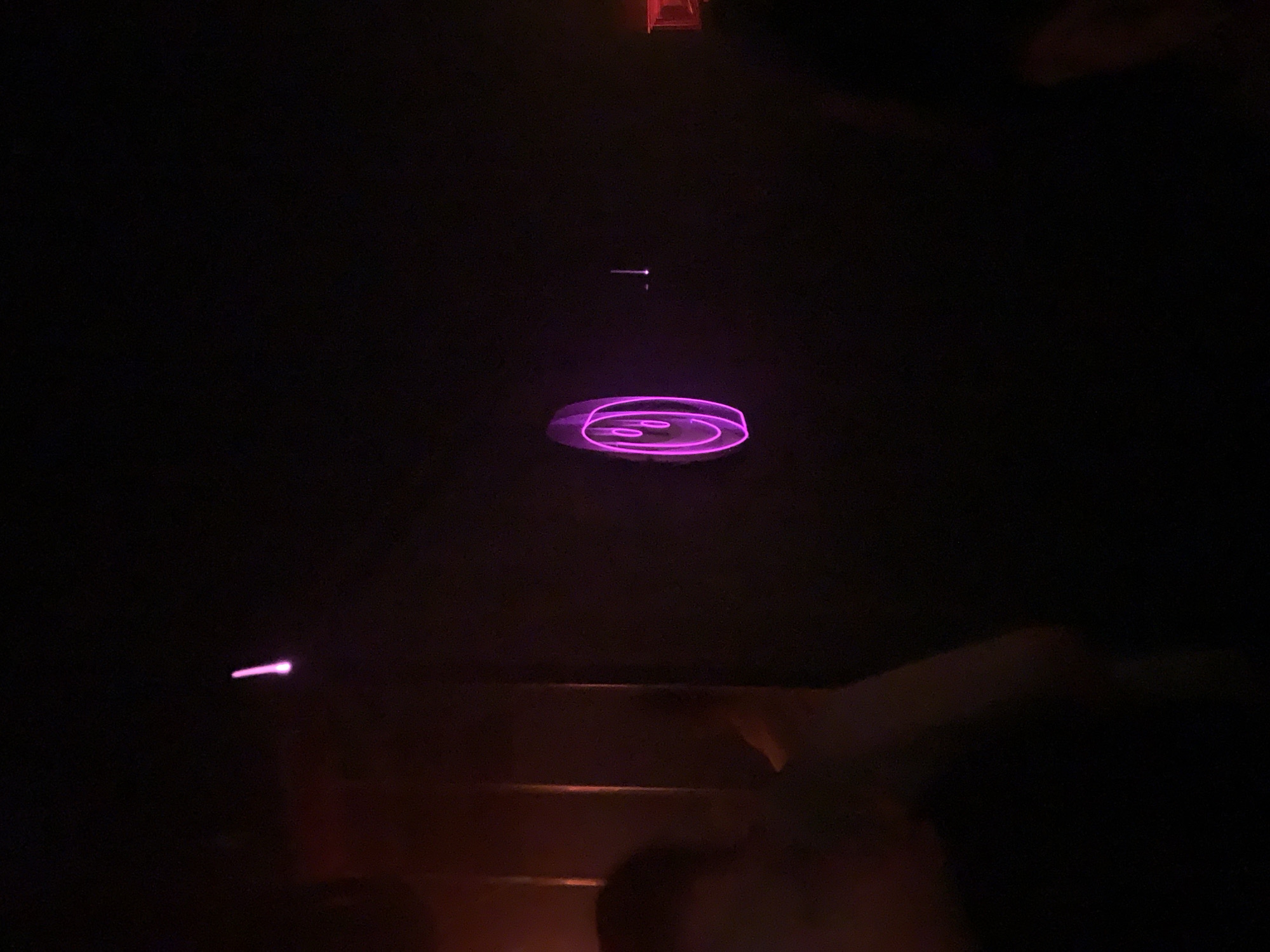 The image, taken in very low light, shows a ceiling with a faint outline of overhead structures and a series of glowing purple projections. The central projection is a series of concentric circles, and there are smaller, linear projections above and to the lower left of it. It is not possible to establish the location in Tel-Aviv, Israel.