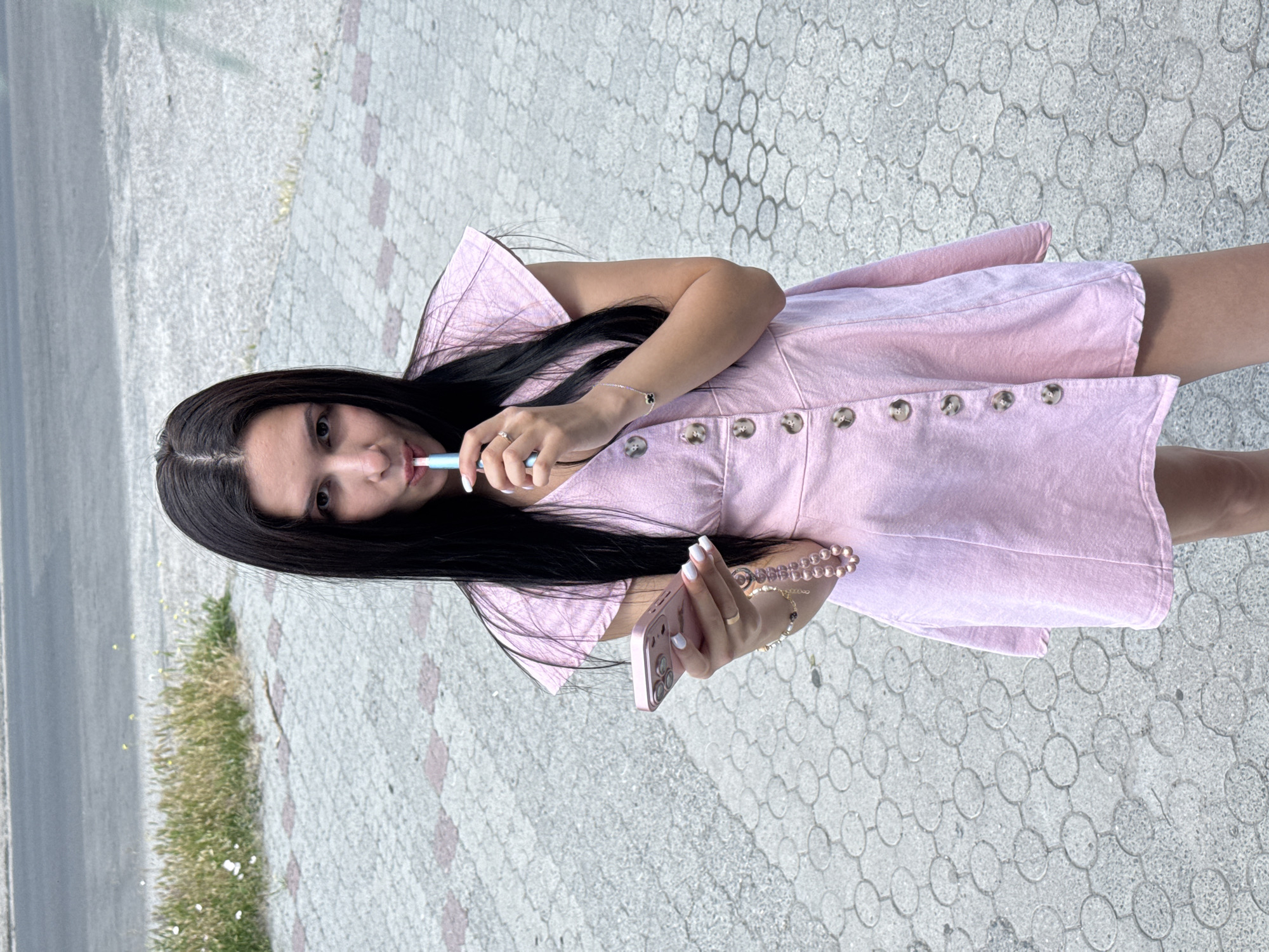 A medium shot shows a woman with long, dark hair wearing a pink dress standing on a paved surface. She holds a pink smartphone in her left hand and a light blue vape device to her mouth with her right hand. The woman has a ring on her left index finger and a bracelet on her right wrist. The paved surface is made of interlocking gray pavers with a pattern of concentric circles. In the background, there is a road and a patch of dry grass.