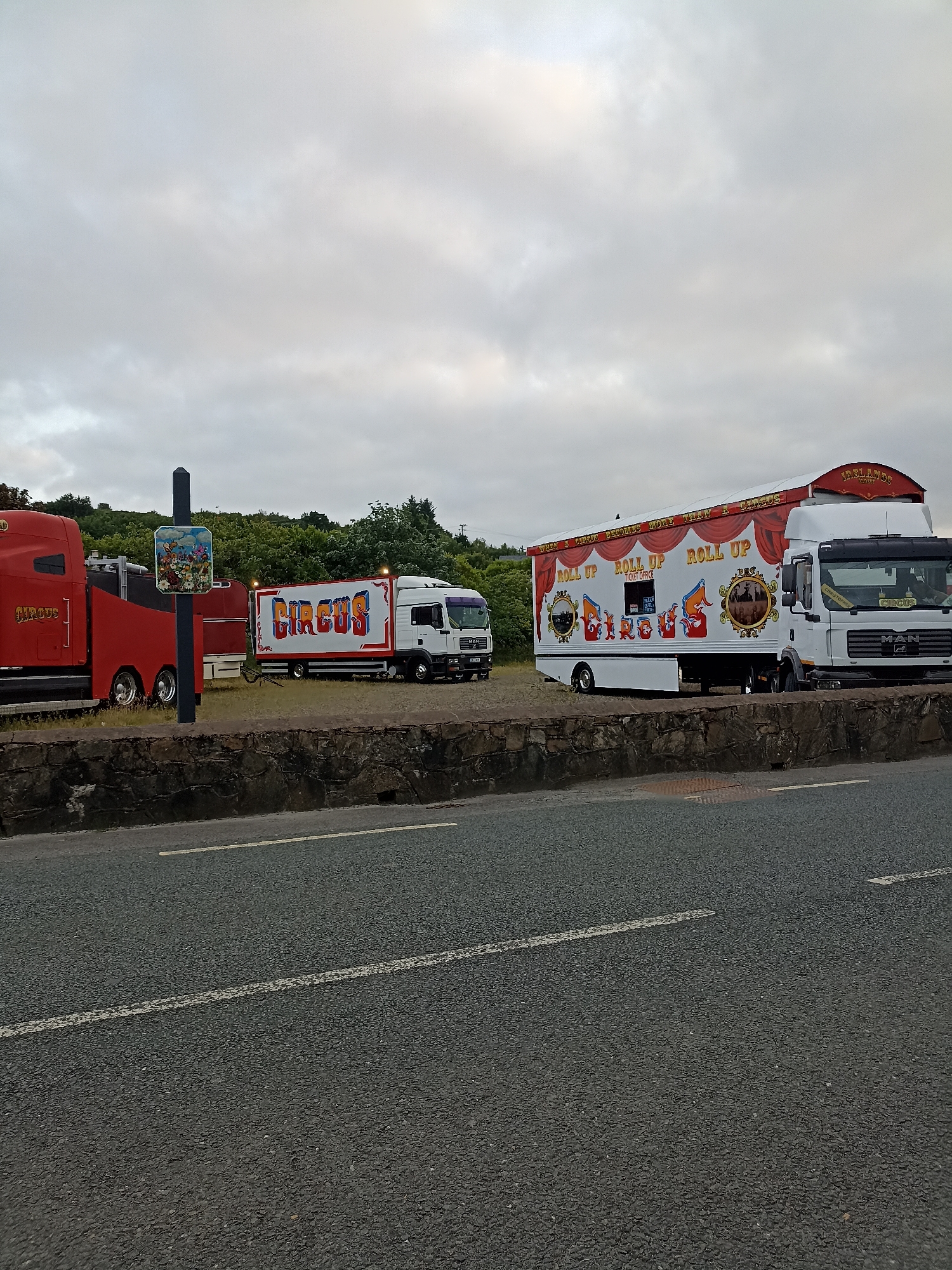 In Annagry, Ireland, a circus appears to be preparing for an event. Two large trucks, one red and one white, are parked on a grassy area behind a stone wall. The red truck has the word "CIRCUS" written on its side in gold lettering. The white truck is larger and more elaborately decorated, also bearing the word "CIRCUS" in large, colorful letters. This truck also has several circular images on its side, and the words "ROLL UP" repeated multiple times. A small sign featuring cartoon characters is mounted on a pole between the two trucks. The sky is overcast, and trees and greenery are visible in the background. A paved road with white lines runs in the foreground of the image.