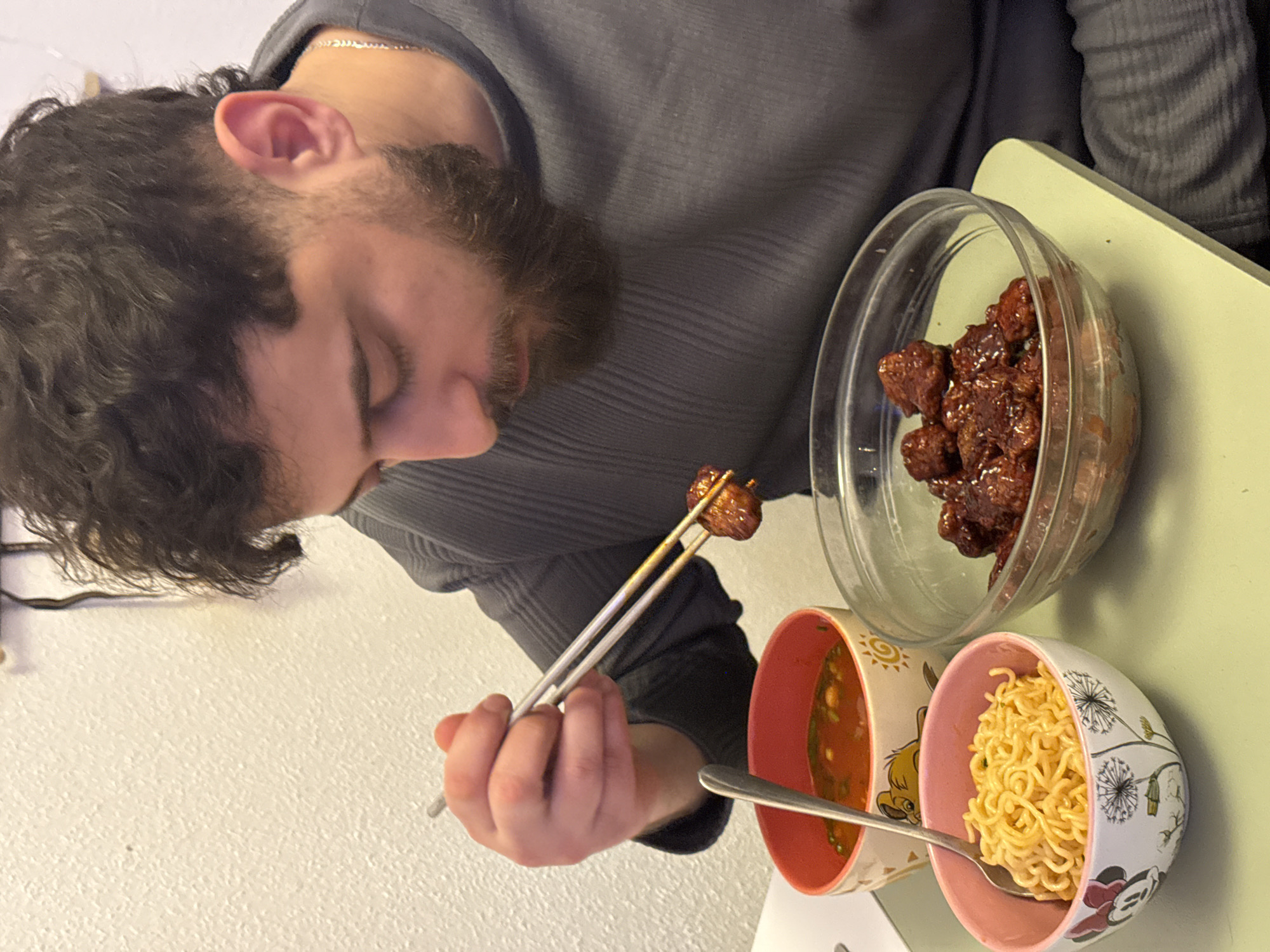 An adult male with dark, curly hair, a faded haircut, and a full beard is depicted seated indoors in Strasbourg, France, consuming a meal. He wears a ribbed, dark grey long-sleeved shirt and a silver neck chain. His head is angled downwards, observing the food. His right hand operates a pair of metallic chopsticks, gripping a single piece of dark reddish-brown, glazed meat extracted from a clear glass bowl. Positioned on a light green surface are two additional food containers: a pink-rimmed white bowl featuring a Minnie Mouse motif, filled with yellow instant noodles and containing a silver spoon; and a red-rimmed white bowl displaying a Lion King motif, holding a red-orange soup also containing a silver spoon. The background consists of a textured, off-white wall.