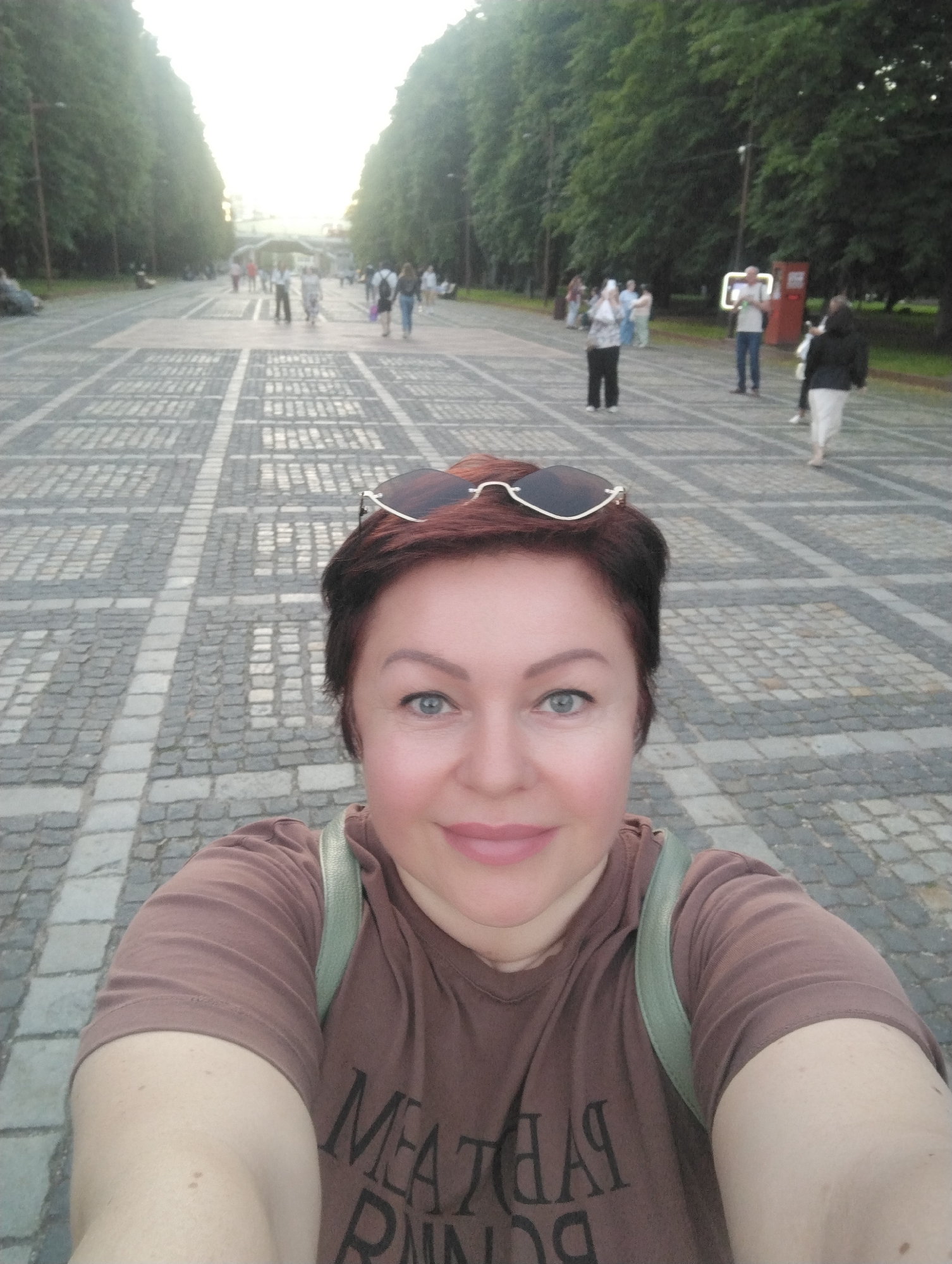 The image is a selfie taken outdoors on a paved walkway lined with trees on both sides. The walkway is paved with rectangular stones in a geometric pattern. In the background, several people are visible, walking and standing. One person is holding up a mobile phone, possibly taking a photo. Sunlight is visible in the upper left portion of the image, suggesting it is daytime. The woman in the foreground, who is taking the selfie, is wearing a brown t-shirt and sunglasses perched on her head. The t-shirt has text printed on it. She has short, dark hair and is looking at the camera with a slight smile. The location is likely a public park or pedestrian area in Kaliningrad, Russia.