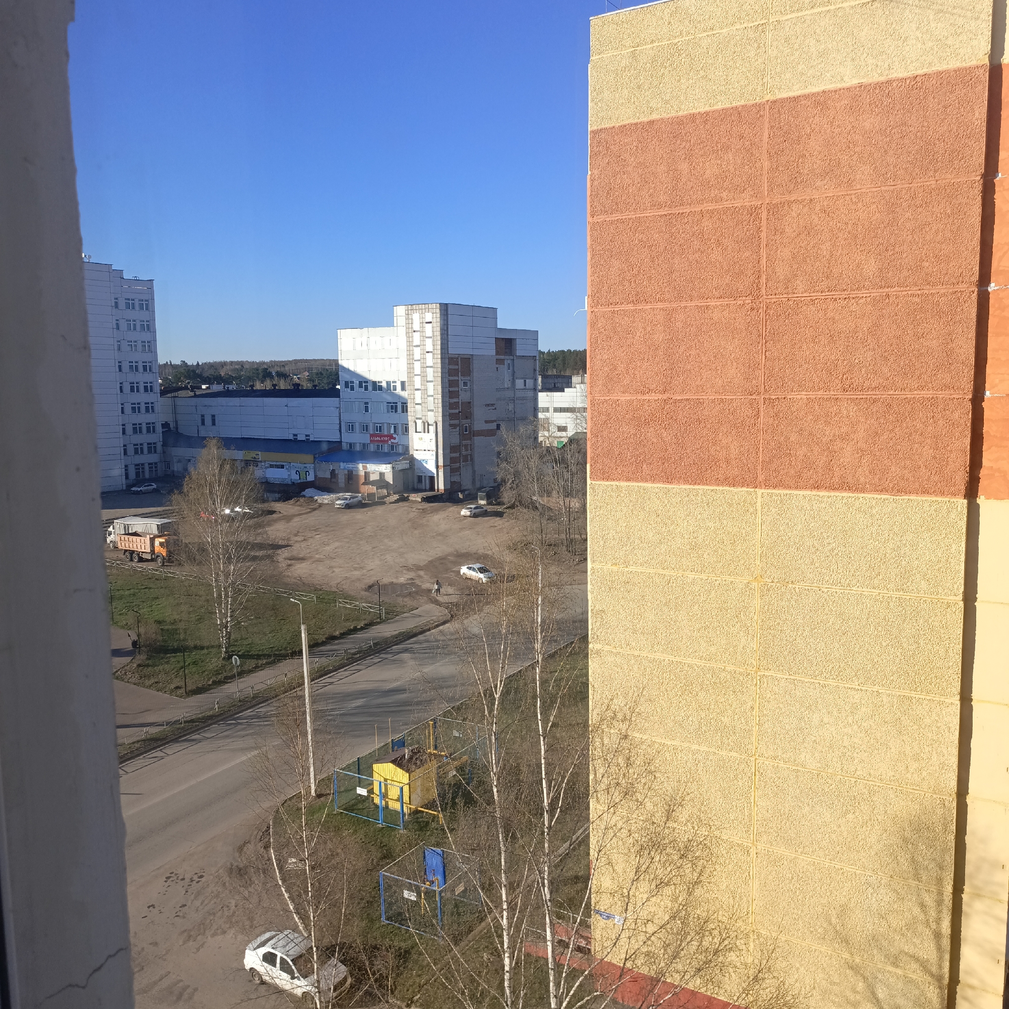 The image shows a view from a high vantage point, possibly a window, looking out over a city scene. Buildings dominate the view, with a large apartment building on the right side of the frame featuring a textured facade in shades of tan and orange. Other buildings, likely commercial or apartment complexes, are visible in the distance to the left, in shades of white and grey.
Below, a road and parking area can be seen, with several parked cars and possibly a pedestrian walking across the lot. Trees, mostly bare of leaves, are scattered throughout the scene, suggesting it is early spring or late autumn. A small fenced-in utility structure painted yellow and blue is near the bottom center of the image. The sky is clear and blue.