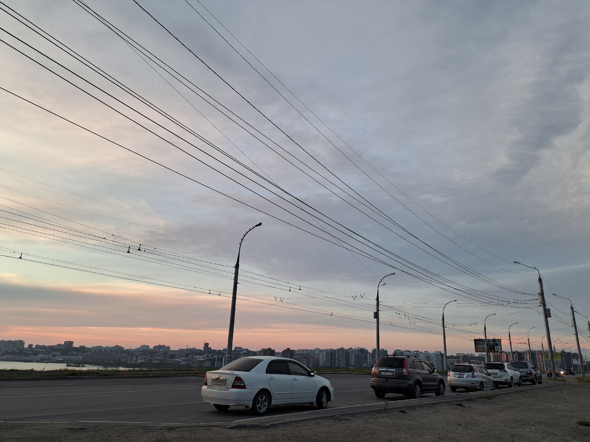 The image depicts a street scene in Irkutsk, Russia, at dusk. Several cars are parked or driving along a road, with a city skyline visible in the background across a body of water. Overhead, a complex network of power lines crisscrosses the sky, illuminated by the fading light of the setting sun, which casts a soft pink and orange glow on the clouds. Streetlights line the road, and the overall atmosphere suggests the transition from day to night in an urban environment.
