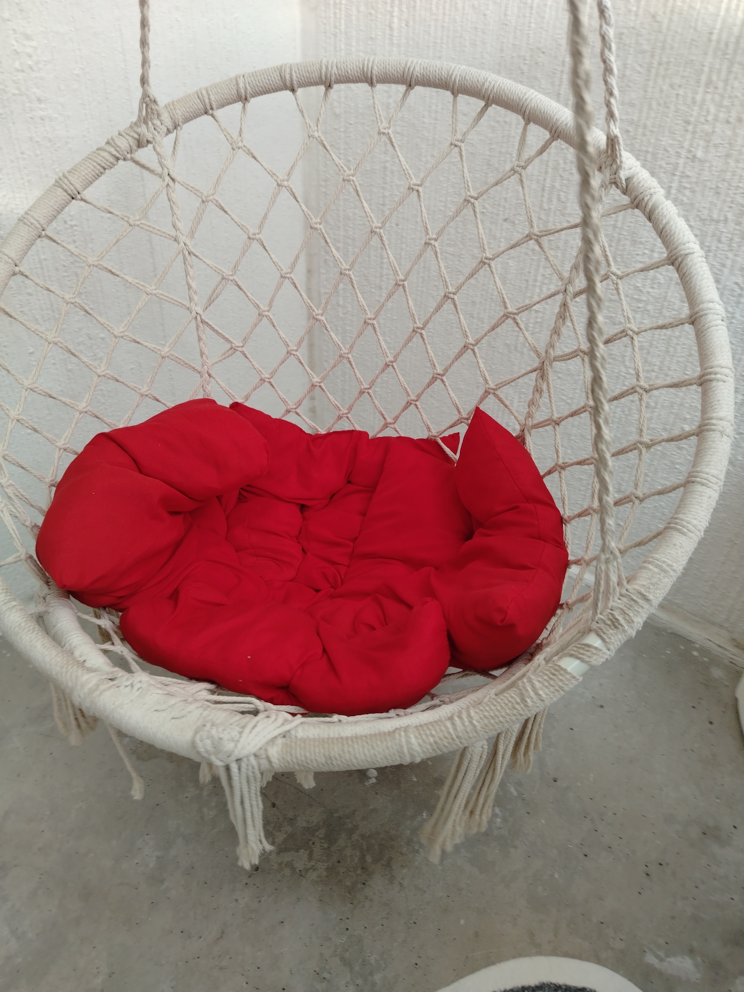 The image displays an unoccupied white macrame hanging chair situated against a textured white wall. The chair features a circular metal frame entirely wrapped in off-white rope, with a diamond-patterned macrame net forming the back and seat. Two thick, braided ropes extend upwards from the top portion of the chair, indicating its suspension mechanism. A bright red, segmented cushion is placed within the chair, appearing soft and conforming to the seat's shape. The floor beneath the chair is a grey, granular surface, consistent with concrete, showing minor dark flecks. A small, circular object with a speckled black and white pattern is partially visible on the floor in the bottom right corner. The scene is static, captured in ambient lighting, and is located in Смалявічы, Belarus.