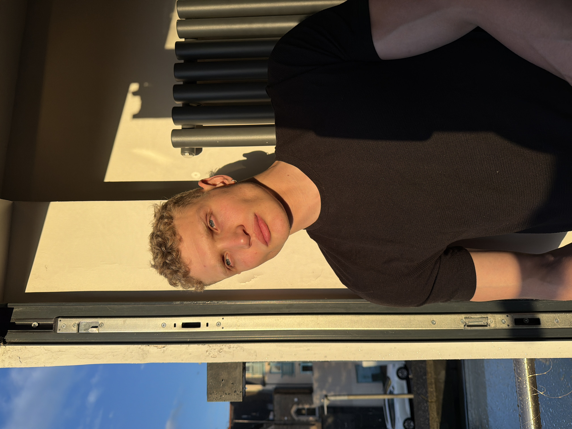 A medium shot depicts a light-skinned person with short, curly, light brown hair and blue eyes. They wear a black long-sleeved shirt. The person is positioned in front of a window, which shows a view of buildings, cars, and a blue sky, indicating an urban location. The image suggests a city setting, potentially London, United Kingdom, due to the architectural style and urban landscape visible through the window. A radiator is partially visible behind the person.