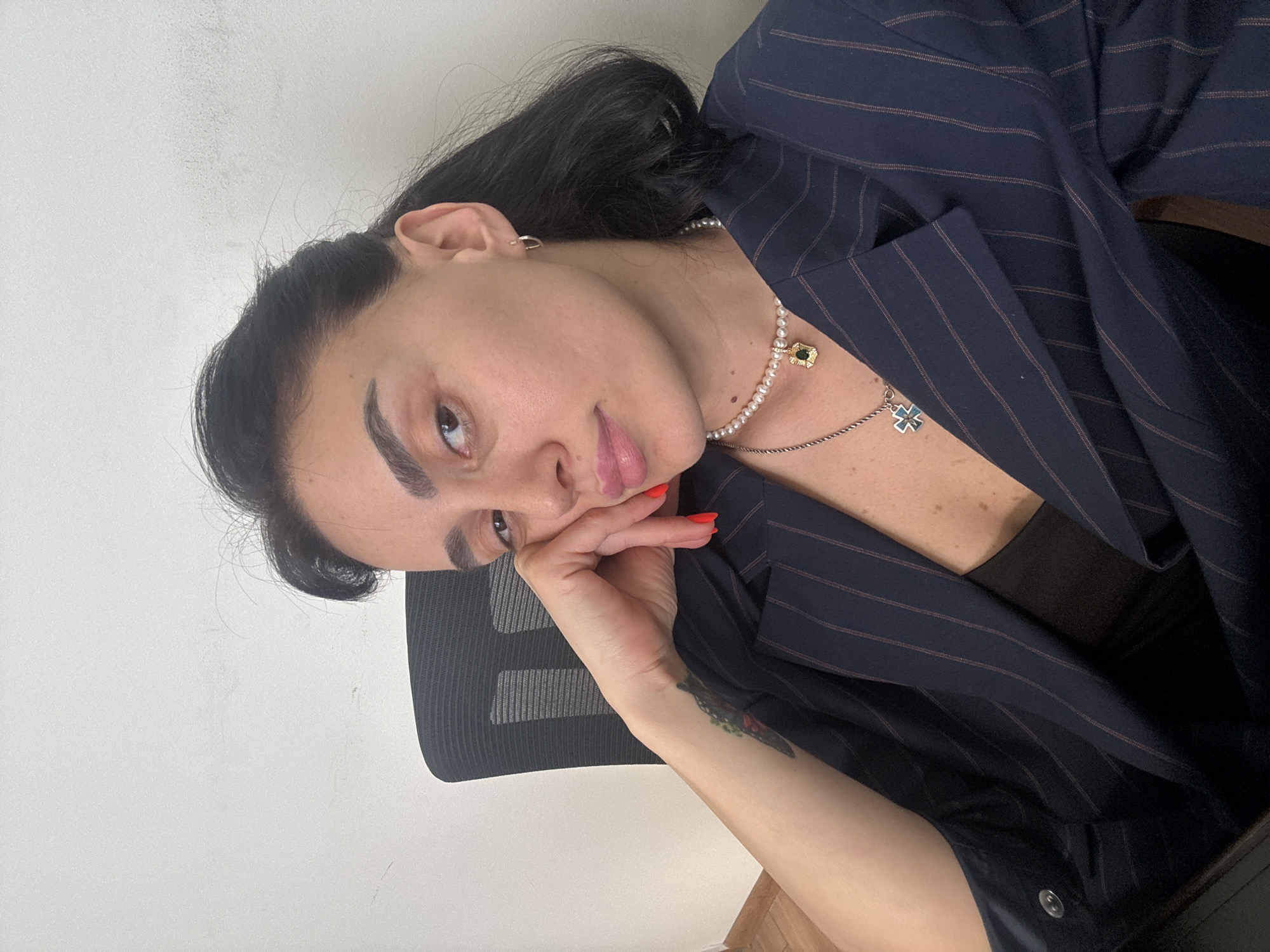 A close-up, vertically oriented portrait features a young adult, likely a woman, with dark hair, prominent dark eyebrows, and light brown eyes. Her complexion is light to medium, and she wears light pink lip gloss and bright red nail polish on her visible hand. She is dressed in a dark blue blazer with thin, reddish-brown vertical pinstripes, worn over a black top.
She accessorizes with two necklaces: a pearl choker featuring a gold-colored pendant with a green gem, and a longer silver-colored chain carrying a distinctive blue and silver cross-shaped pendant. A small silver-colored hoop earring is visible in her right ear. Her left hand, with red nail polish, rests gently against her left cheek. A dark, possibly floral or plant-themed, tattoo is visible on her inner left forearm/wrist.
The setting is indoors, with a plain, light-colored wall (likely white or off-white) serving as the background. A partial view of a black office-style chair is seen behind her on the left side of the frame. The lighting is even, suggesting an indoor environment or soft daylight. The subject looks directly at the camera with a neutral to slightly pensive or relaxed expression. There is no visible text in the image. The provided context indicates the location is Tbilisi, Georgia.