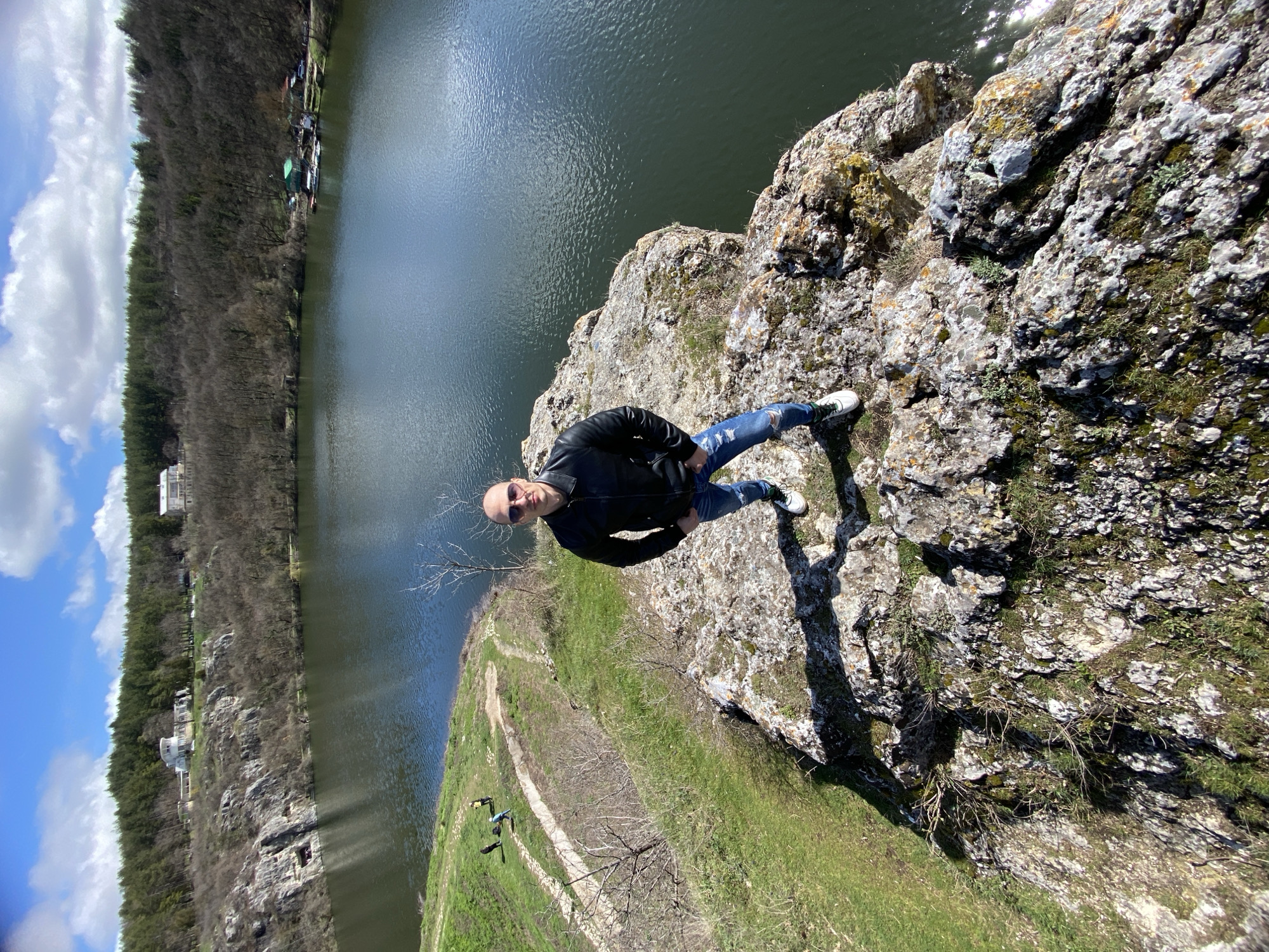 The image captures an elevated view of a natural landscape within Kayluka Park, Pleven, Bulgaria. In the foreground, a bald adult male, wearing dark sunglasses, a black leather jacket, ripped blue jeans, and white sneakers, stands on a prominent, lichen-covered rocky outcrop. He faces the camera directly, hands inserted into his pockets. To his left and below, a dirt path winds down a verdant grassy slope, upon which three distant individuals are visible. The central and background portions of the image are dominated by a large, dark blue-green body of water, identified as Kayluka Lake, exhibiting surface ripples and reflections. The far bank consists of a steep, rocky cliff covered primarily by leafless deciduous trees and denser coniferous forestation on the upper slopes. On the distant hillside, two prominent white buildings are visible: one on the left with a distinctive modern design featuring a rounded upper section, and another on the right with a more conventional structure. At the water's edge on the right, a green-roofed building complex with small boats docked nearby suggests a recreational facility. The sky is clear blue with scattered white cumulus clouds, indicating a bright, sunny day.
