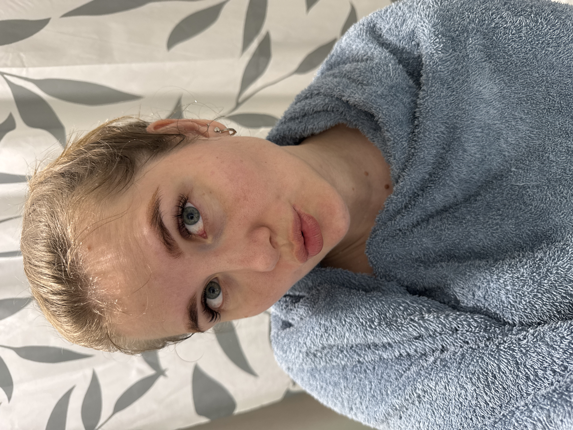 A woman is posing in front of a backdrop in the location of Ферма, Russia. She is wearing a light blue towel. Her hair is blonde. The backdrop is white with gray leaf patterns.