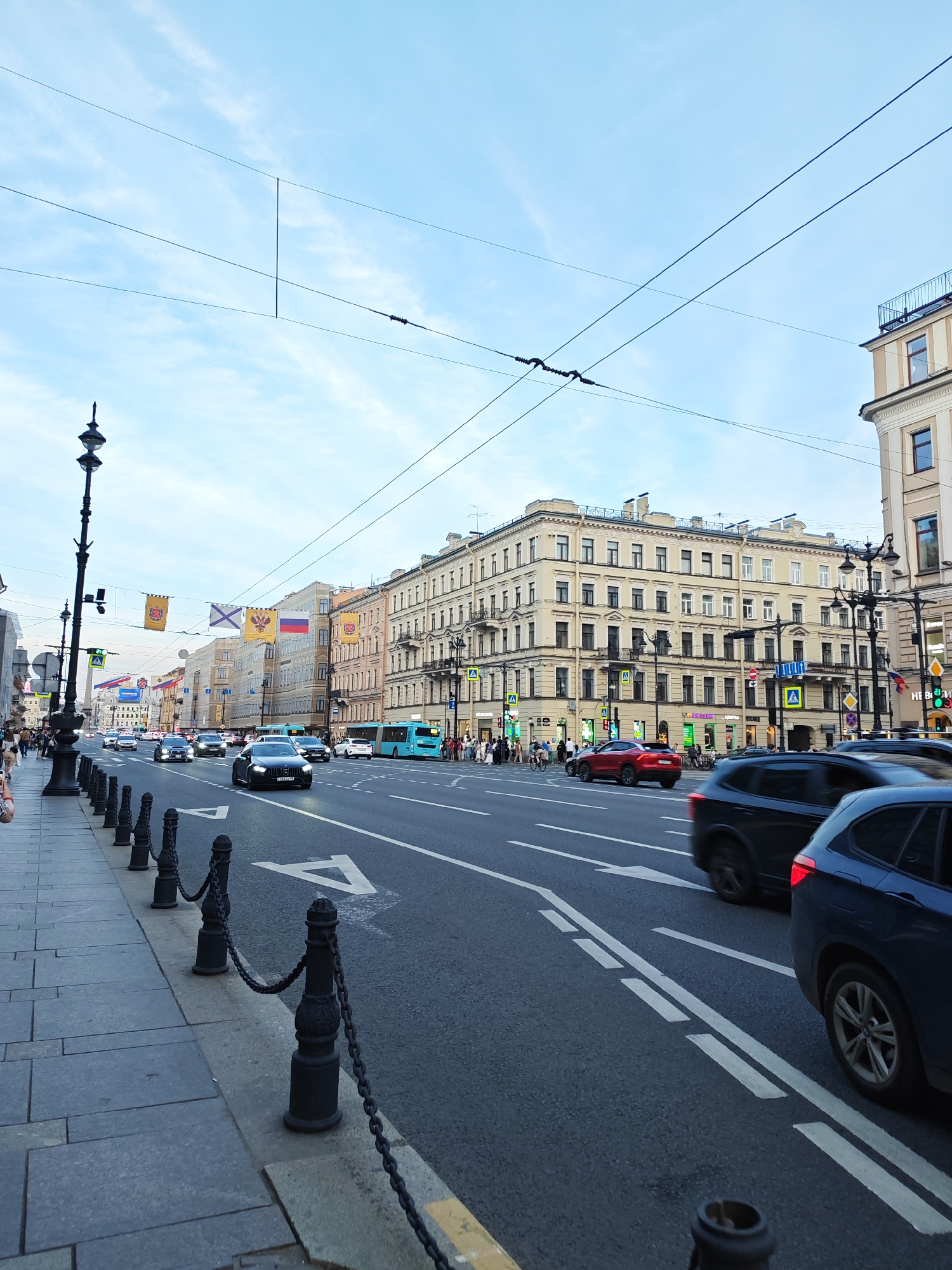 This image depicts a wide, bustling urban street, identified as Nevsky Prospekt in Saint Petersburg, Russia. The perspective is from the left sidewalk, looking down the multi-lane thoroughfare under a light blue sky with scattered white clouds, suggesting late afternoon or early evening.
The street is paved with asphalt, featuring multiple lanes demarcated by white markings, including a double solid line, dashed lines, and prominent white arrows indicating traffic direction. Overhead wires, characteristic of trolleybus infrastructure, span across the street. Numerous vehicles occupy the lanes; a dark-colored sedan with its headlights on is prominent in the mid-left lane, facing away from the viewer. Other vehicles include a red SUV and several dark-colored SUVs, some exhibiting motion blur. A light blue trolleybus is visible further down the street on the right side.
Along both sides of the street, multi-story historical buildings with classical architectural elements dominate the skyline. These structures feature light-colored facades, numerous windows, and decorative details such as cornices and balconies. On the left sidewalk, a tall, ornate black lamppost stands prominently, and a series of black bollards connected by chains separate the pedestrian area from the roadway. The sidewalks are paved with grey stone slabs.
Numerous pedestrians are visible on the sidewalks on both sides of the street and gathered at intersections. Overhead, across the street, decorative banners and flags are strung. These include yellow banners with emblems, and three distinct Russian flags: one displaying the double-headed imperial eagle, another the St. Andrew's Cross (naval flag), and the third the modern Russian tricolor. Traffic lights are visible at intersections, with a green light illuminated on the right side of the frame.
