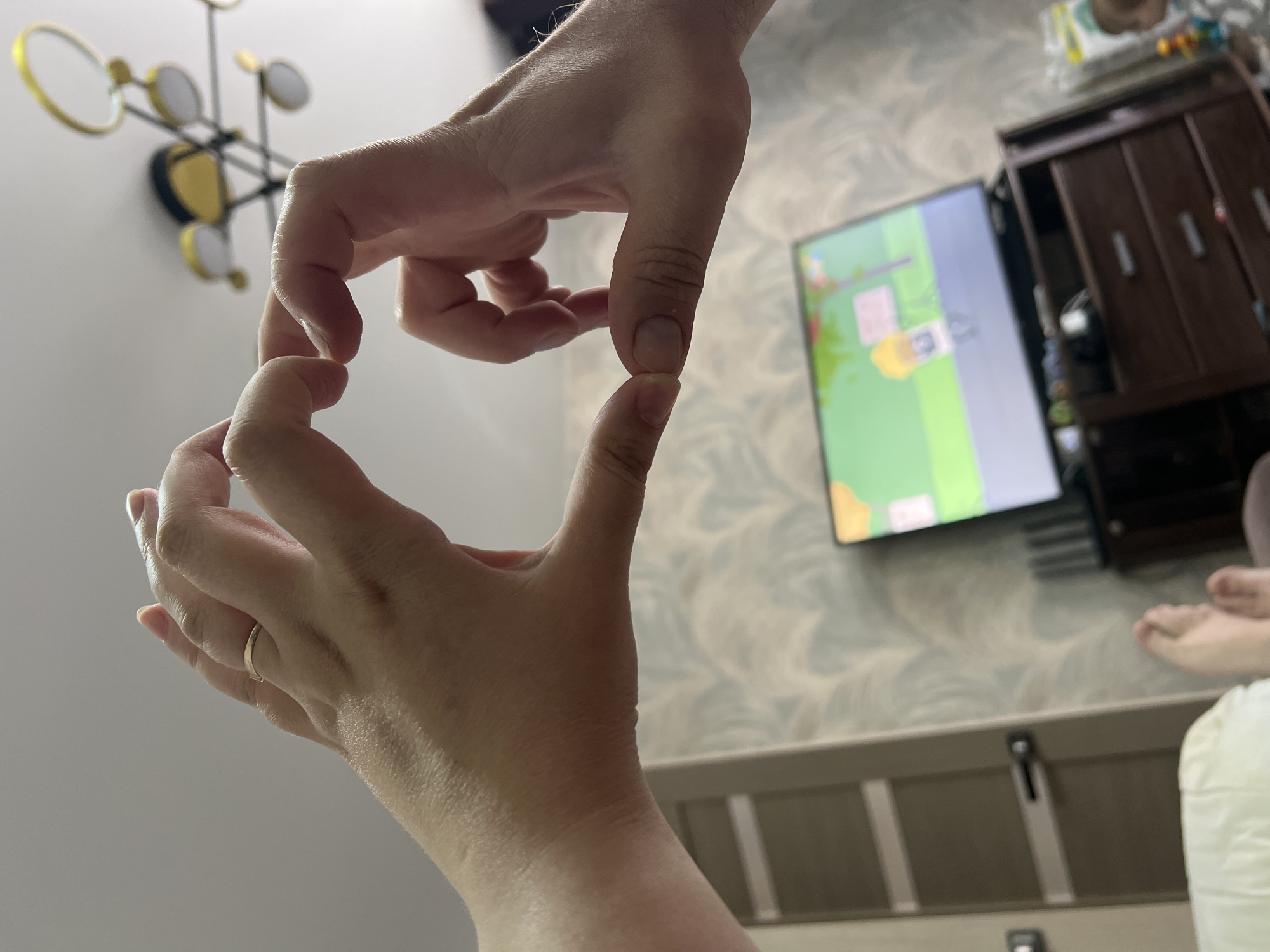 The image shows two hands forming a heart shape, presumably for a photo. A television is visible in the background, along with a cabinet and a portion of bedding. The location is Noviy, Russia.