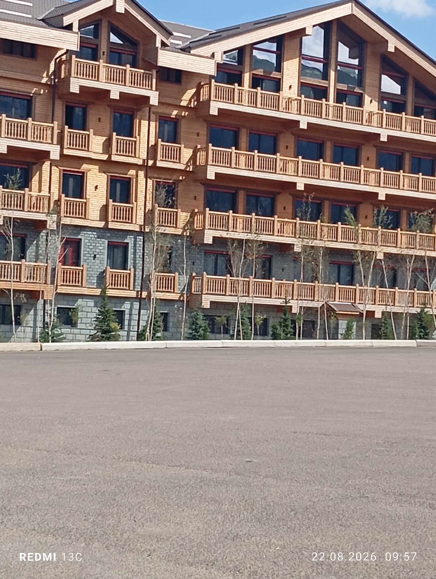 The image shows the exterior of a multi-story building with a wooden facade and balconies, located at the Amirsoy ski resort in Uzbekistan. The building has a modern design with large windows and a sloping roof. Trees and evergreen shrubs are visible in front of the building. The foreground is an asphalt-paved area, likely a parking lot or access road. The sky is clear blue with some clouds. The bottom left corner of the image has the text "REDMI 13C" and the bottom right corner has "22.08.2026 09:57".