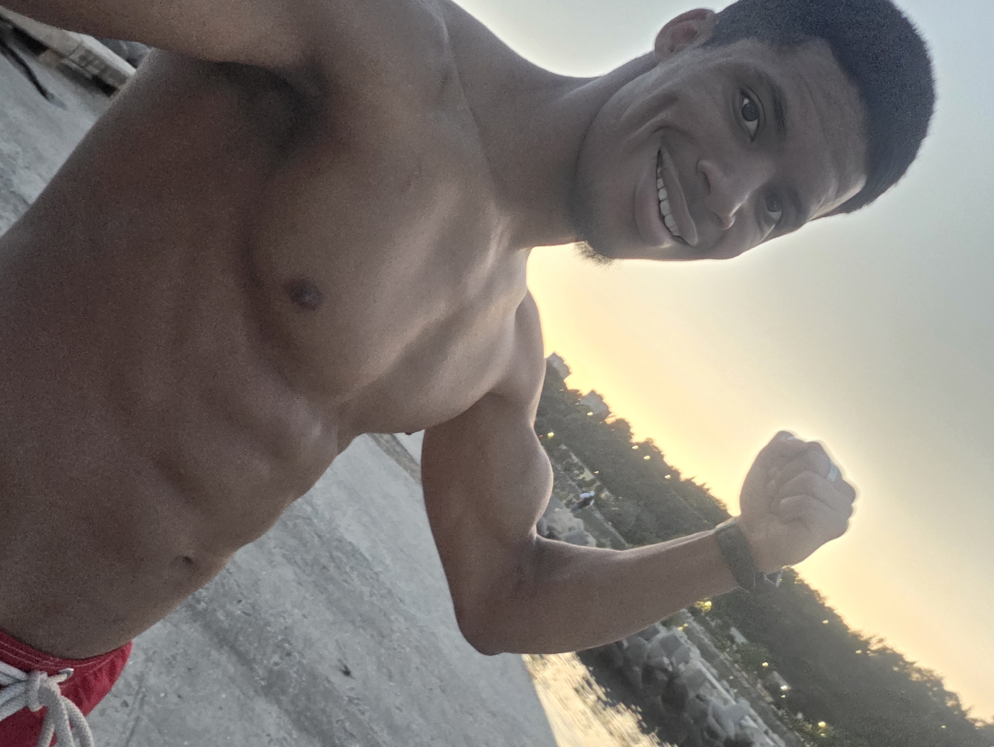 A man with dark skin and short dark hair is posing for a photo outdoors at dusk. He is shirtless, wearing red swim trunks. He is flexing his right bicep and smiling at the camera. The background shows a body of water, rocks, and a tree-lined hillside with lights. The scene appears to be a coastal area. The presence of a small goatee on the man's chin is noted. The sky is a soft yellow and orange, indicating the sun is setting.
