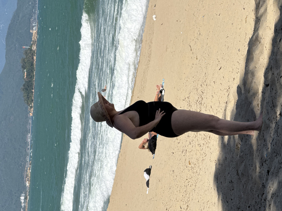 A woman in a black swimsuit and a straw hat is lying on a sandy beach next to the ocean. Her hand rests on her stomach. Other people and items such as clothing are nearby. The beach runs parallel to the ocean, with waves crashing along the shore. In the distance, a coastline with trees and buildings can be seen. A long shadow stretches across the beach.